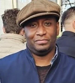 A close-up portrait of a man with a serious expression, wearing a checkered shirt and a dark sweater, against a plain light-colored background.