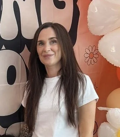 Young man with a beard and mustache wearing a tan cap, black jacket with fur collar, and white shirt, seated indoors in a restaurant or cafe with illuminated street view in the background.