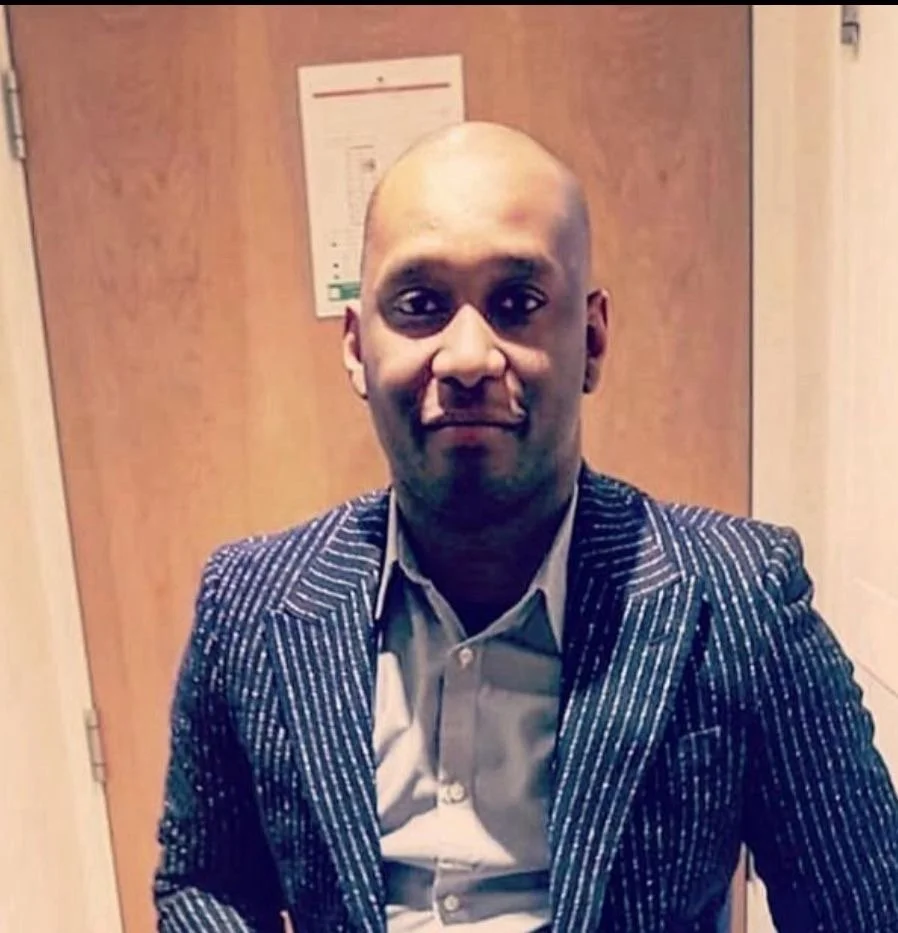 A close-up portrait of a man with a serious expression, wearing a checkered shirt and a dark sweater, against a plain light-colored background.