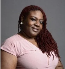 A woman with long, red braids wearing a pink top and pearl earrings, looking at the camera with a slight smile.