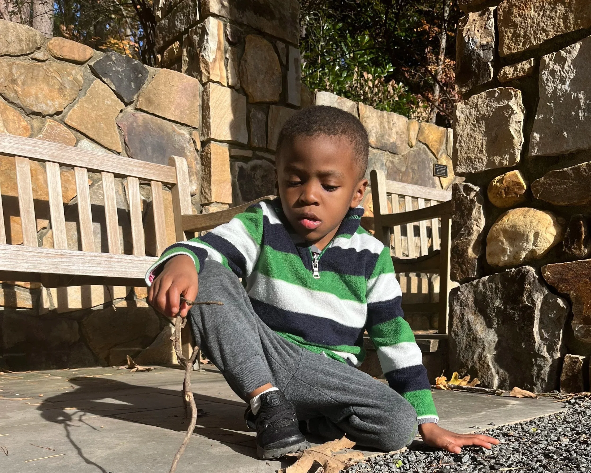 A young boy wearing a green, white, navy, and gray striped sweater and dark gray pants is crouched down outdoors on a paved surface, holding a twig or small stick, with stone benches and a stone wall in the background.