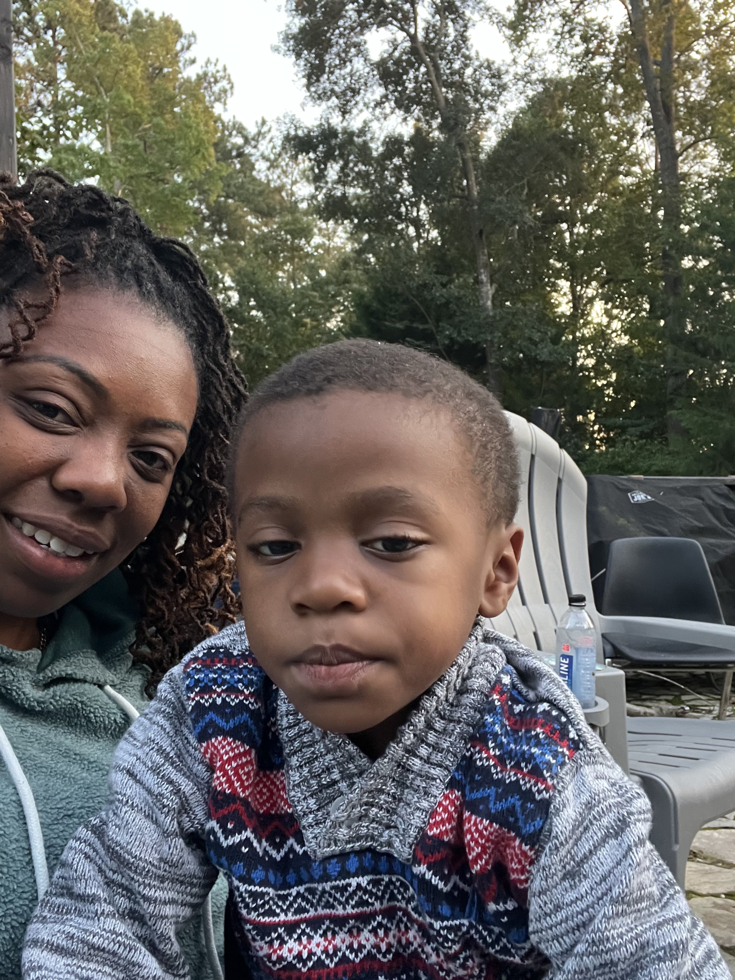 A woman and a young boy sitting outdoors, with trees and outdoor chairs in the background, and a water bottle on a chair next to them.