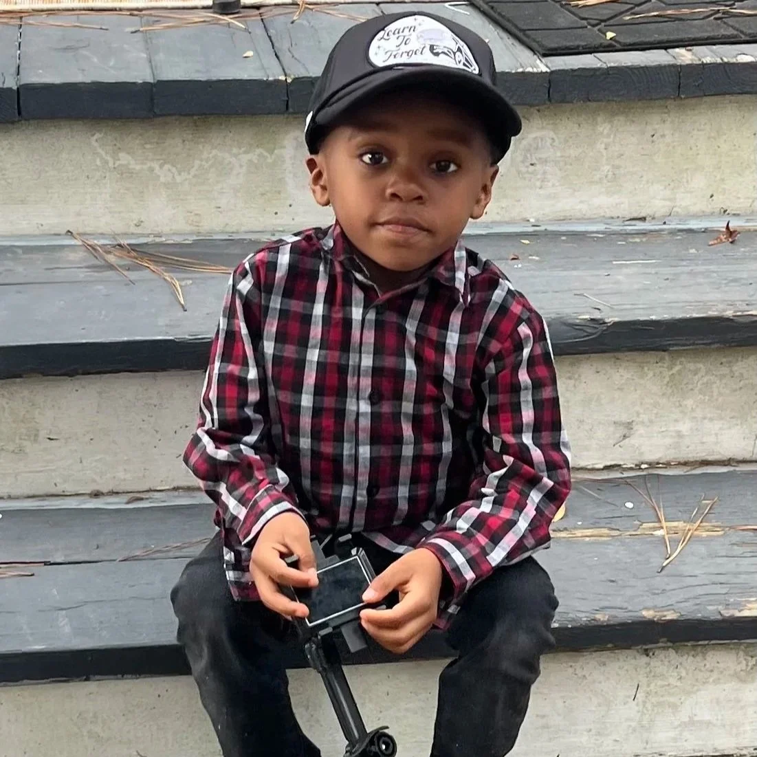 A young boy wearing a black cap with a logo and the words 'Learn To Forged,' a red and black checkered shirt, and dark pants, sitting on outdoor stairs, holding a camera in his hands, with a neutral expression.