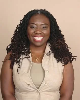 Portrait of a smiling African American woman with curly hair, wearing a beige sleeveless vest and a light-colored top.