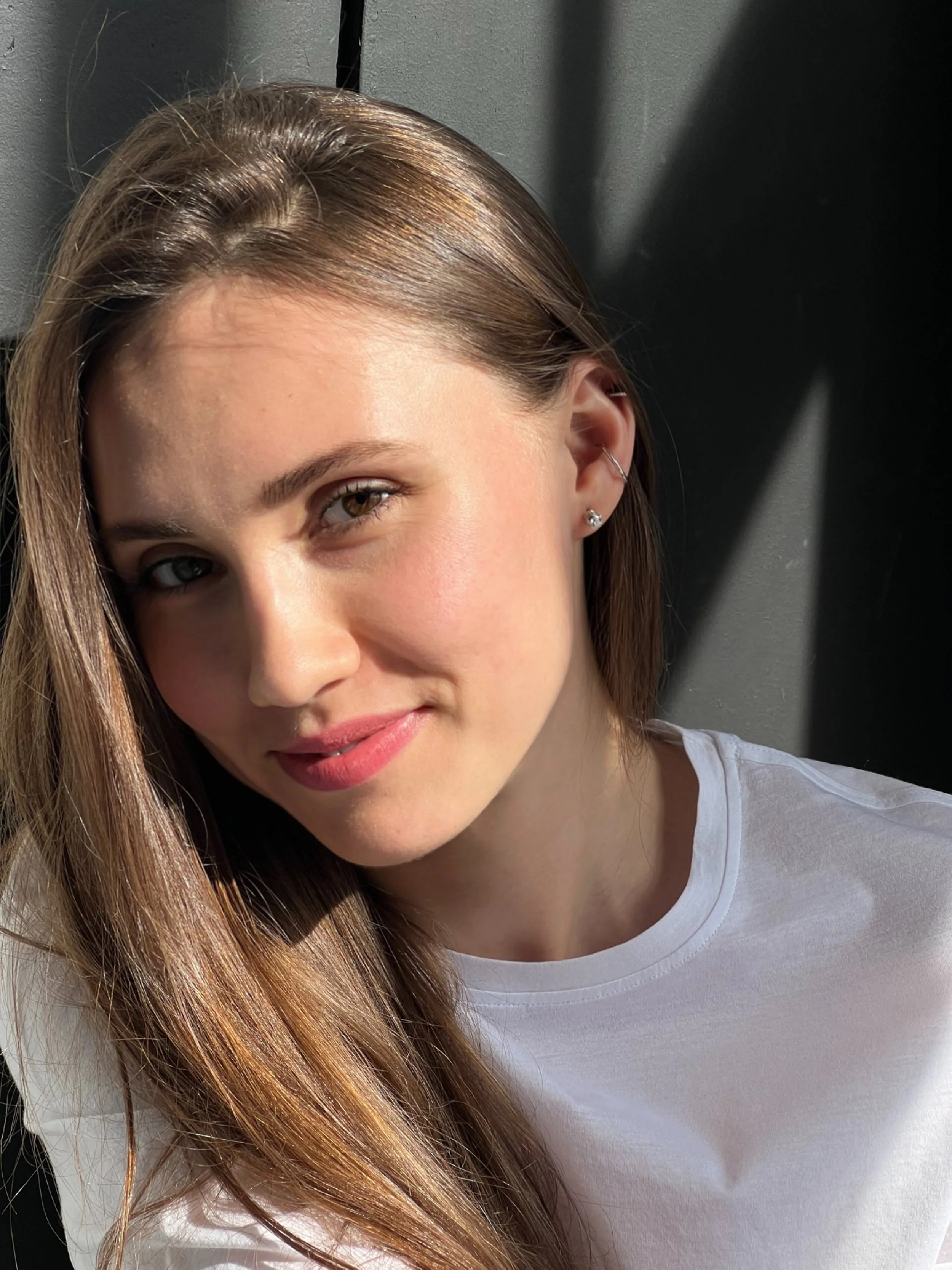 A young woman with long, straight, light brown hair and light skin, wearing a white t-shirt, outdoors in sunlight.