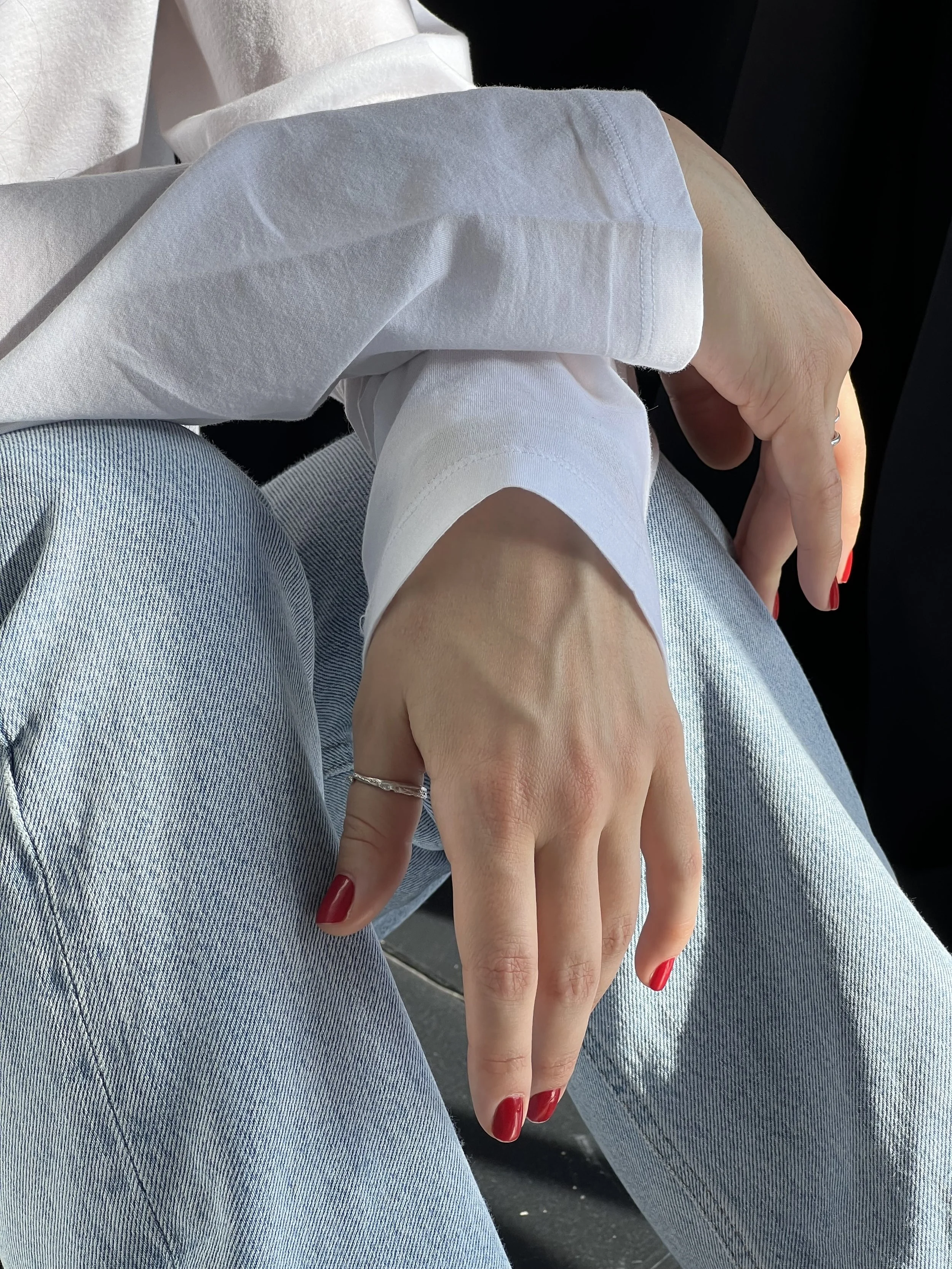 Close-up of a woman's hand with red nails, resting on her knee, wearing light blue jeans and a white shirt with rolled-up sleeves.