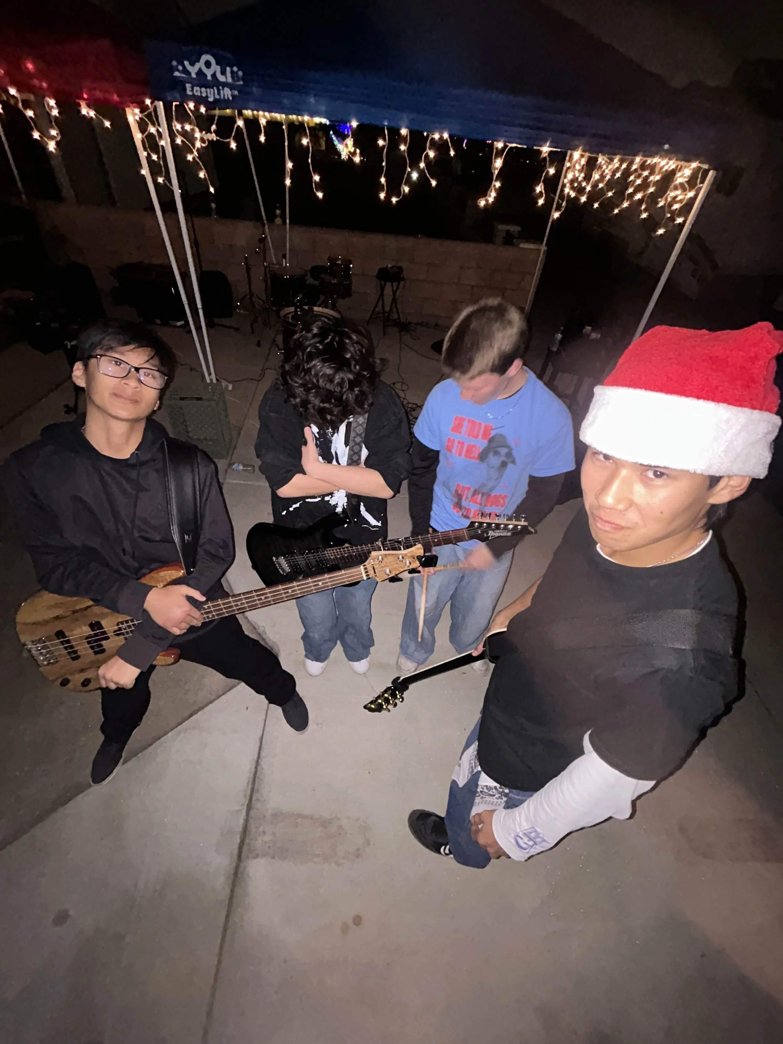 Four young males with guitars standing outdoors at night, Christmas lights overhead, one wearing a Santa hat.