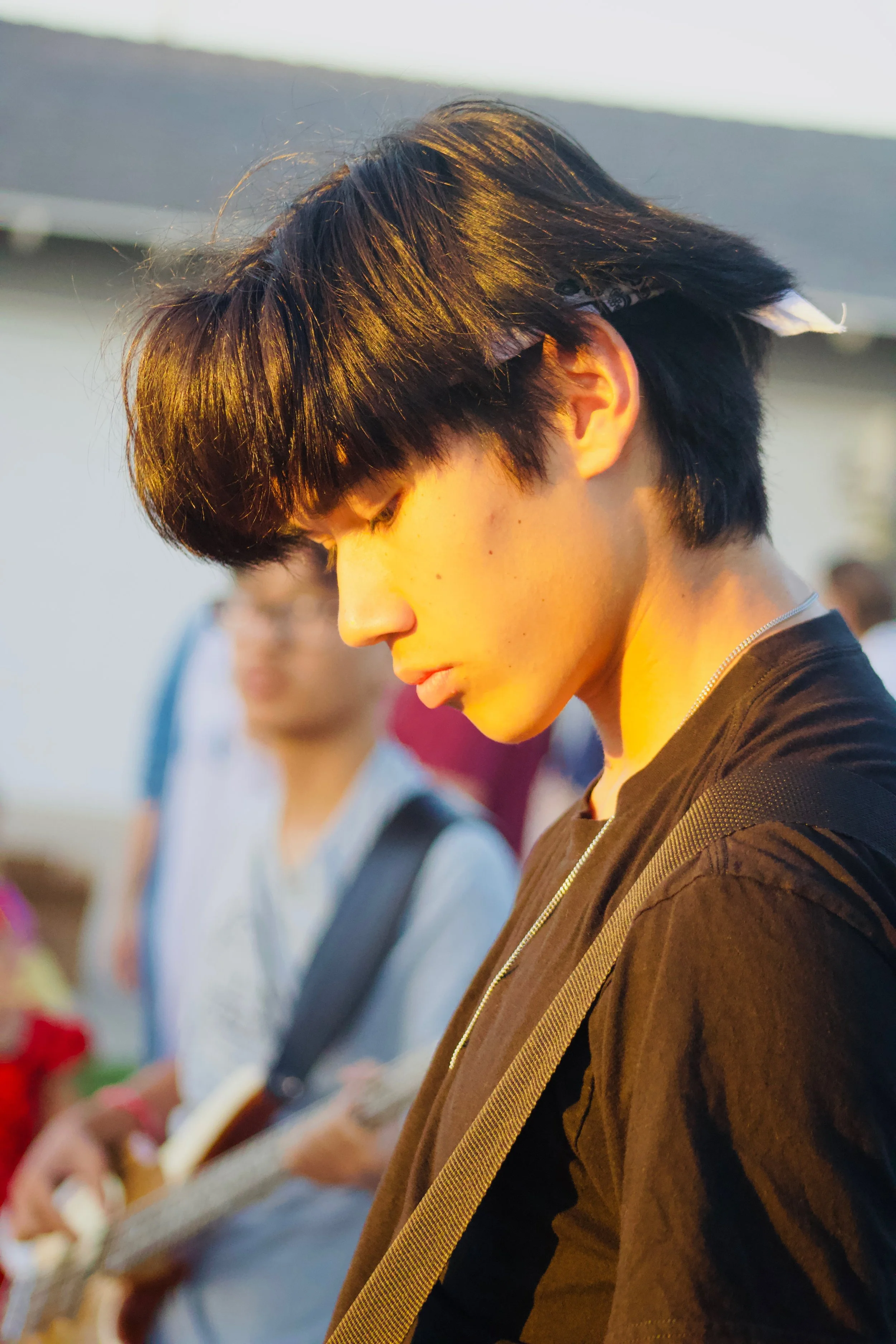 Side profile of a young man with dark hair and a black shirt, with a person playing guitar blurred in the background, outdoors at sunset.