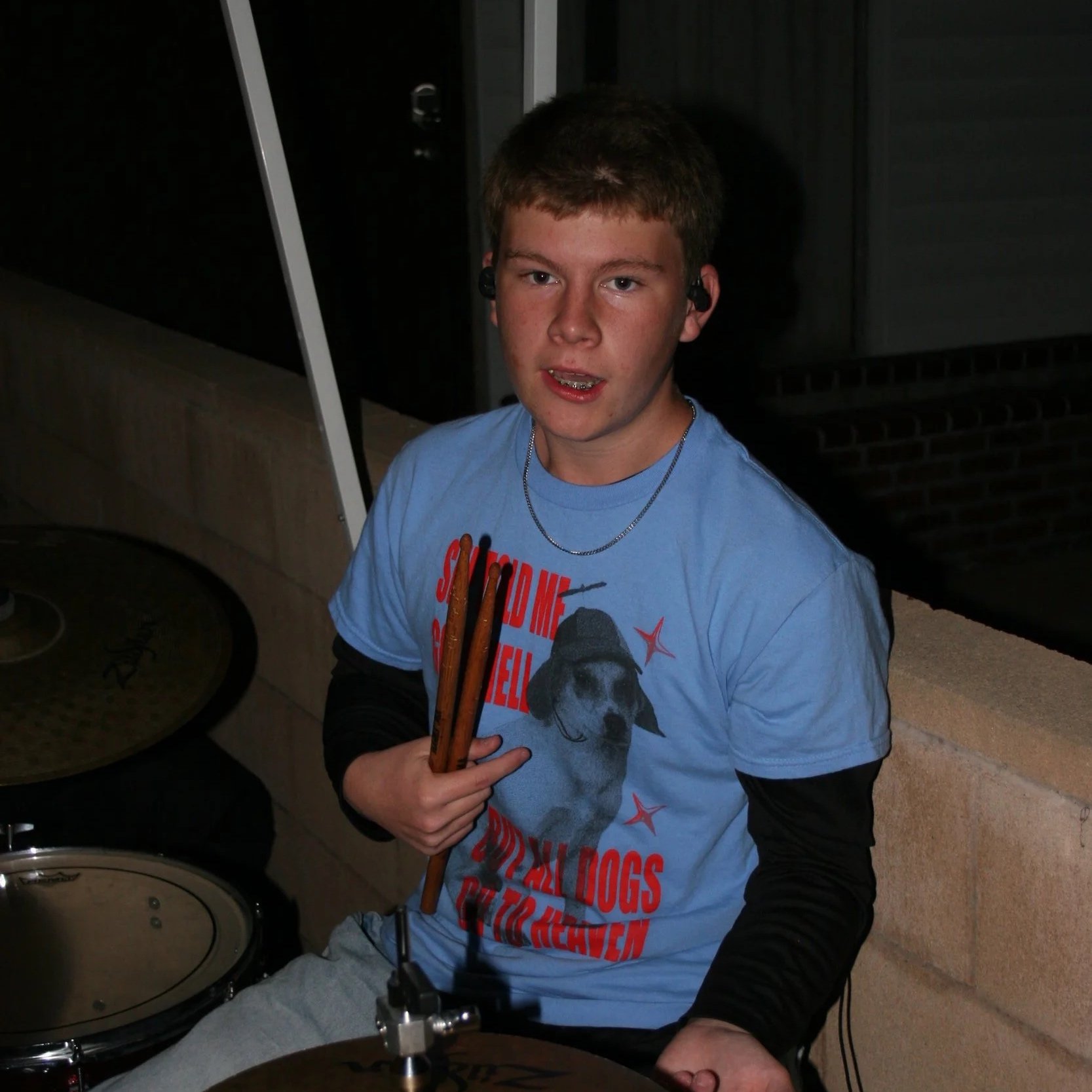 A young man with short brown hair sitting at a drum set, holding drumsticks, wearing a blue t-shirt with dog graphics, and a black long sleeve shirt underneath, in a dimly lit room.
