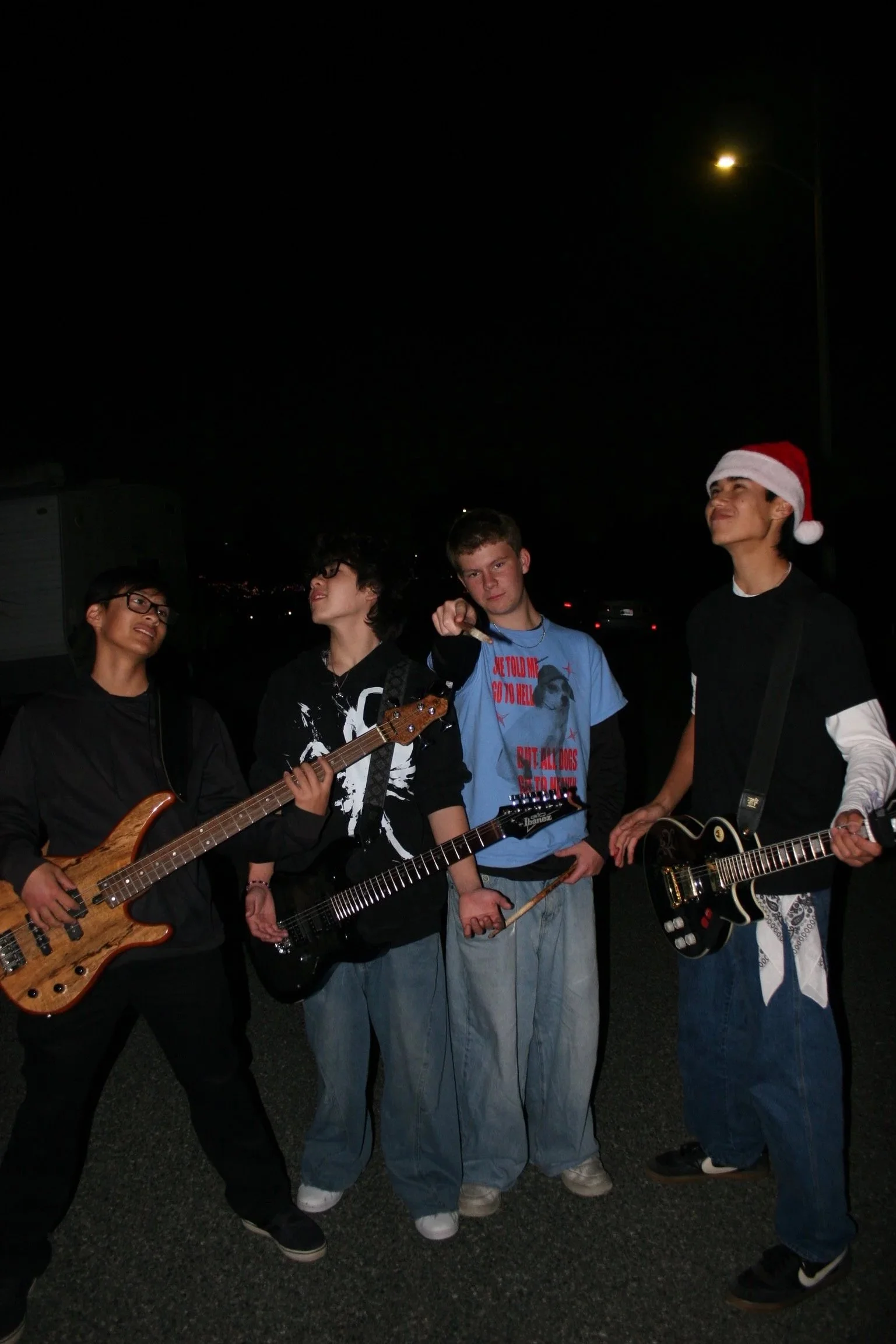 Four young men standing outdoors at night, holding musical instruments; one wears a Santa hat, suggesting a Christmas theme.