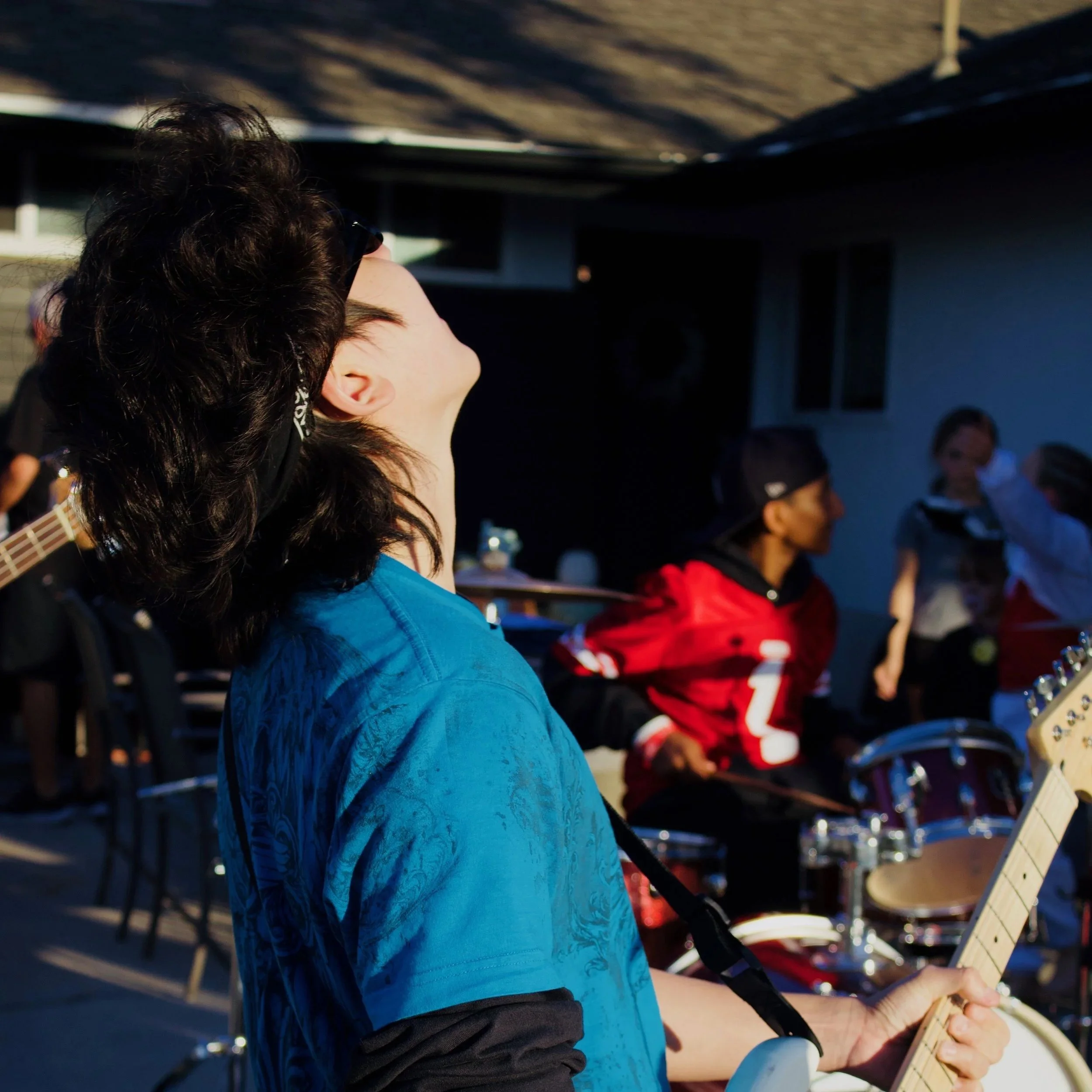 A person playing a guitar with long dark hair, wearing a blue shirt, outdoors with a group of people in the background.