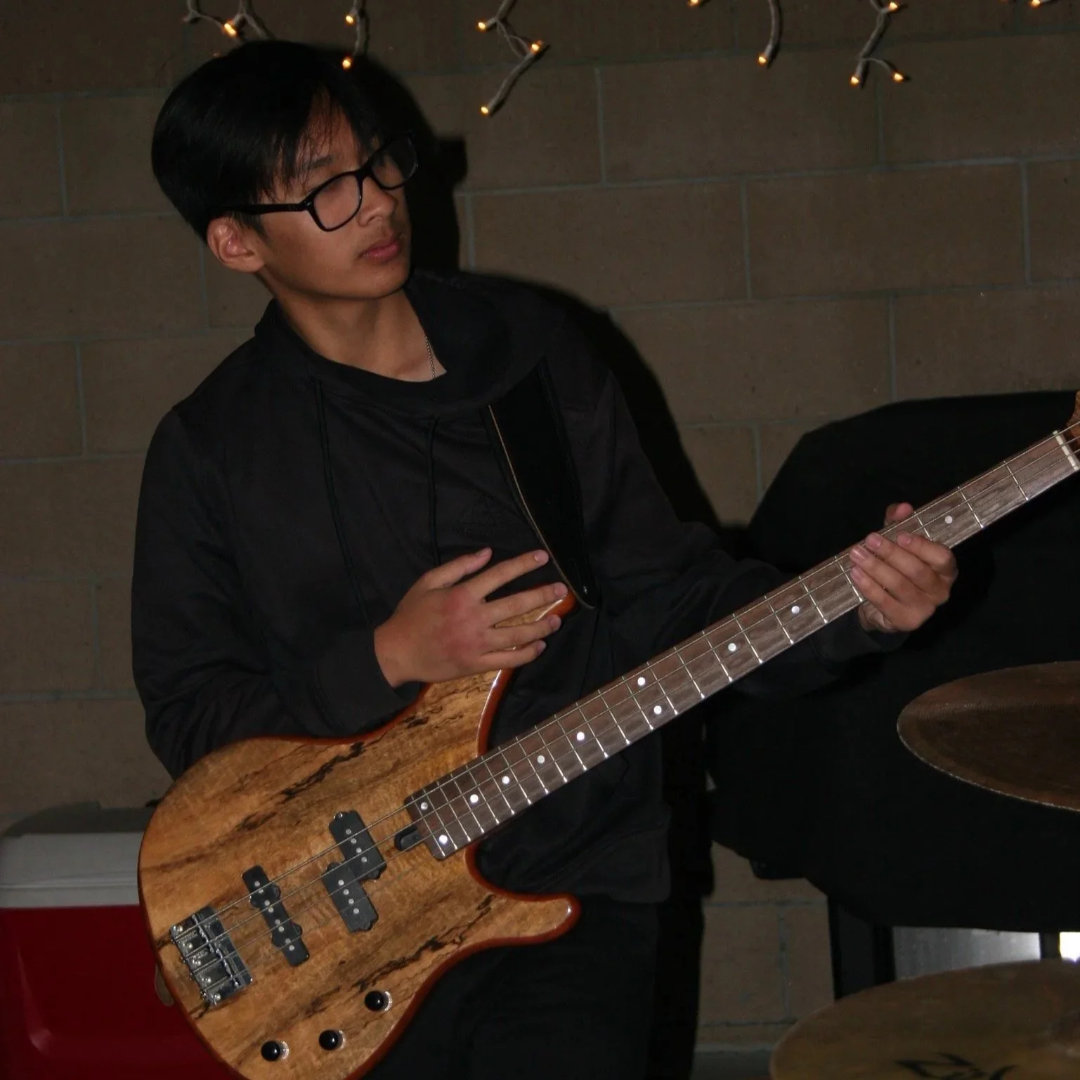 A young man with black hair and glasses playing an electric bass guitar with a natural wood finish indoors, against a brick wall decorated with string lights.
