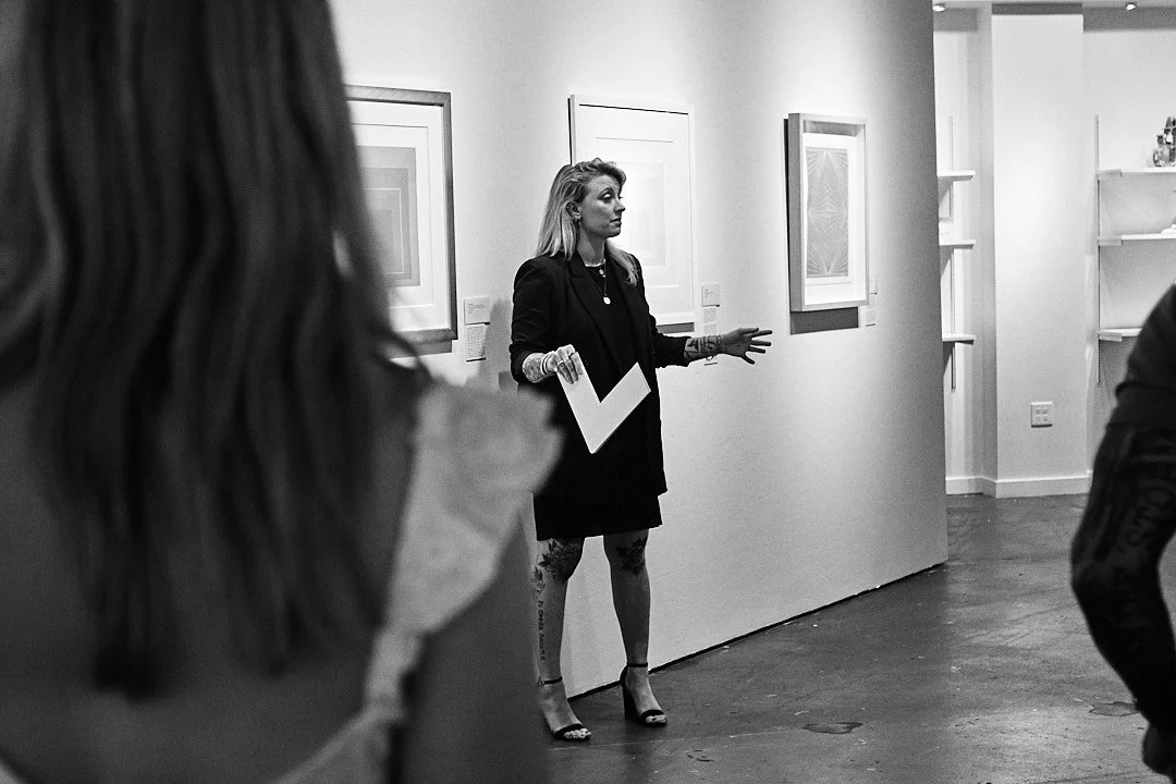 Black and white photo of Allee Beatty speaking to a group of collectors in a gallery, holding a piece of art