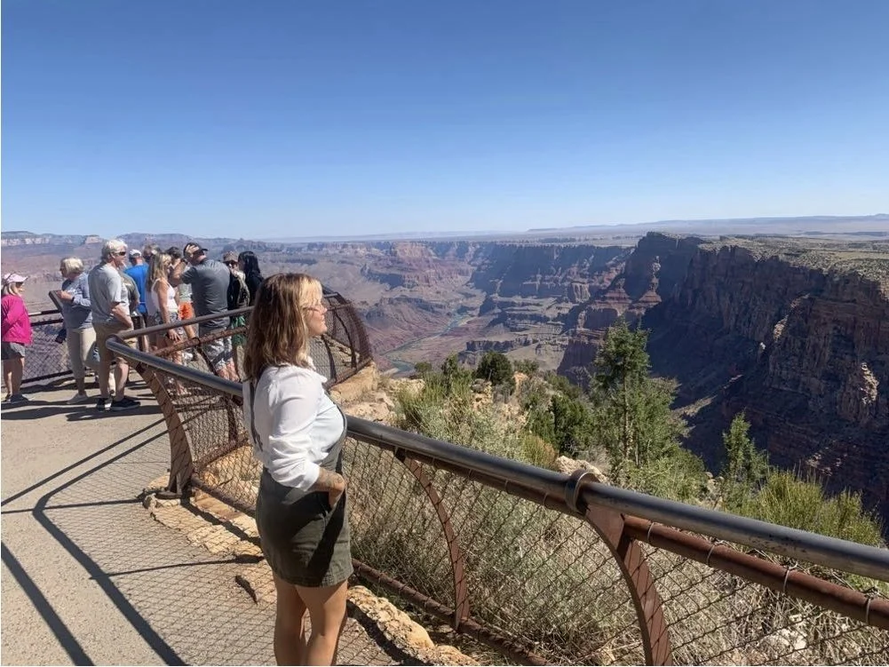 Tourists standing at a viewpoint overlooking the Grand Canyon on a sunny day.