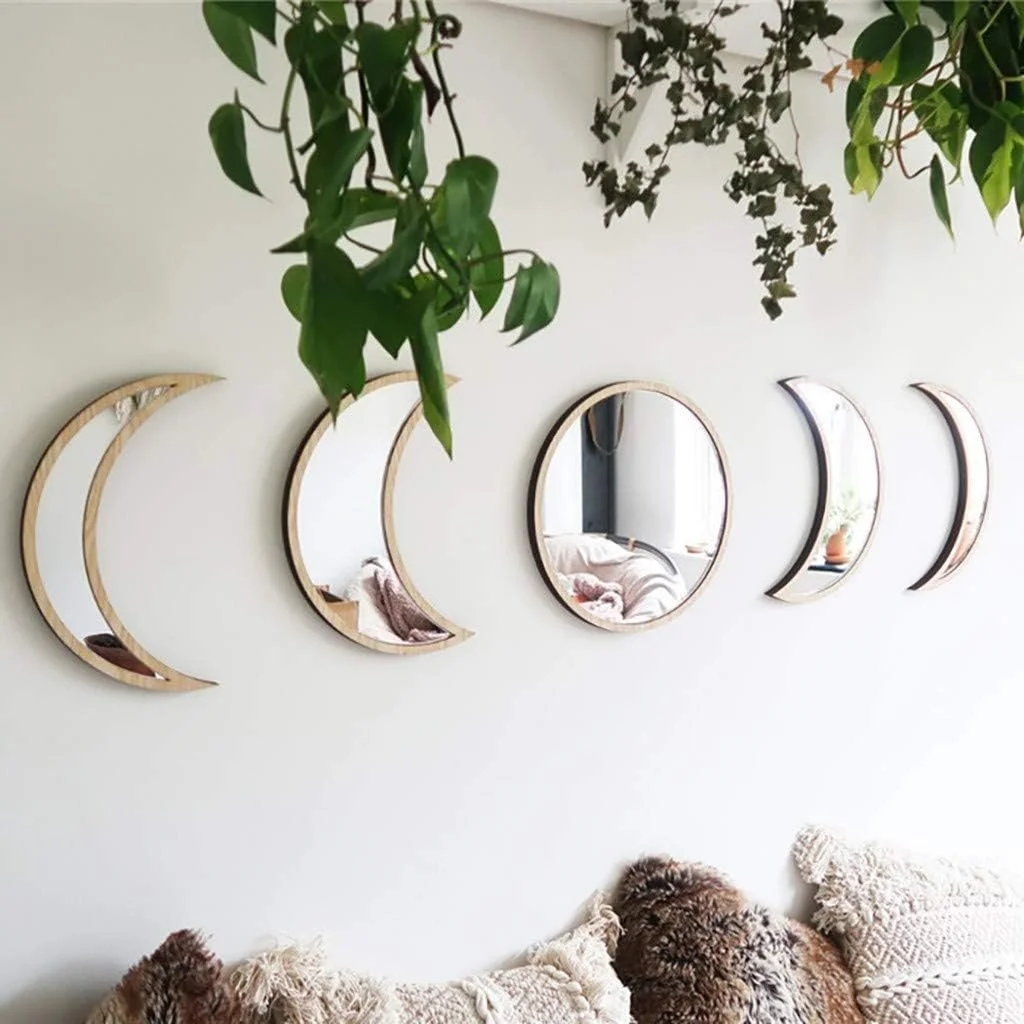 A white wall decorated with six crescent moon and one oval-shaped mirror, with potted plants hanging above, and a cozy couch with plush pillows below.