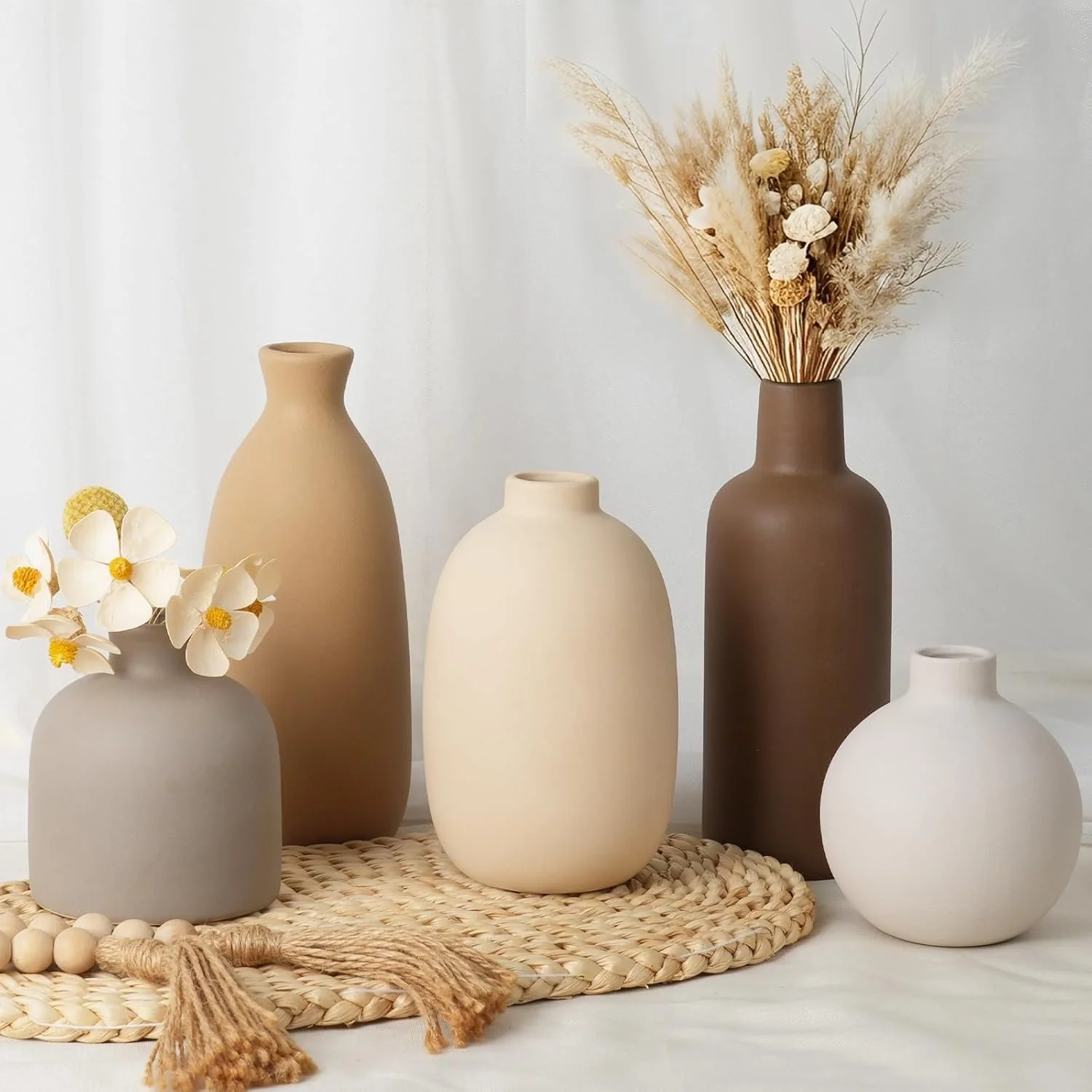 Arrangement of five minimalist ceramic vases, some with dried flowers and pampas grass, on a woven mat with a white background.