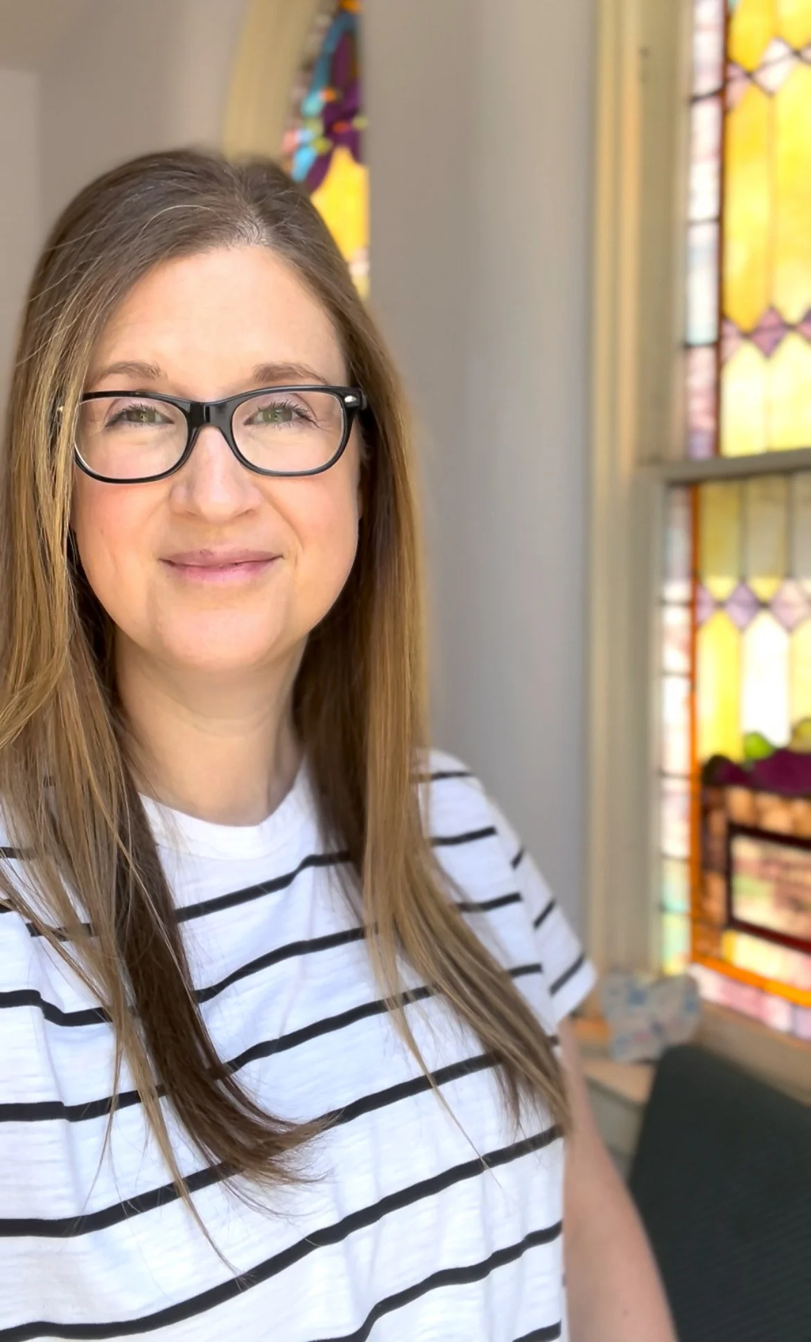 A woman with long brown hair, wearing glasses and a white shirt with black horizontal stripes, standing inside a building with stained glass windows.