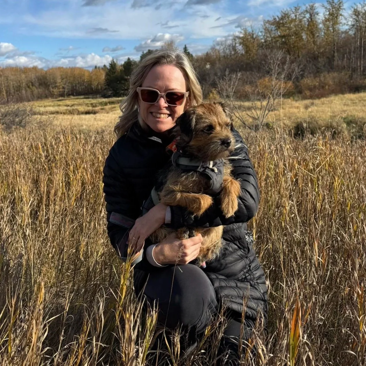 A woman with sunglasses smiling and holding a small brown and black puppy in a field of tall yellow grass, with trees and a partly cloudy sky in the background.