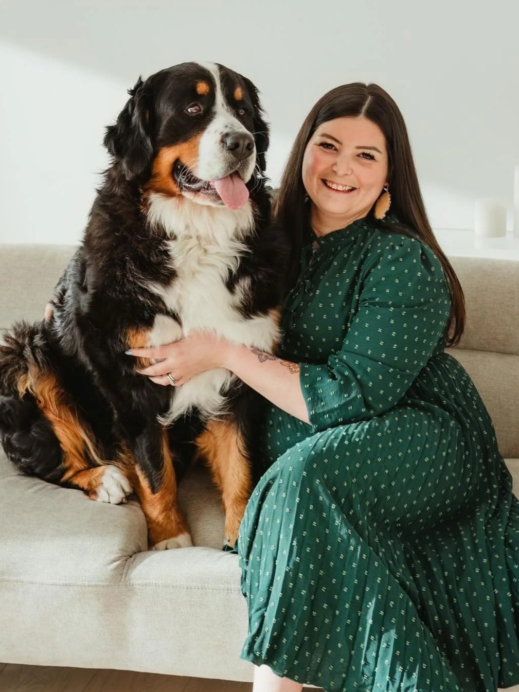A woman with long dark hair, wearing a green dress with small white patterns, sitting on a beige sofa, smiling and holding a large Bernese Mountain Dog.