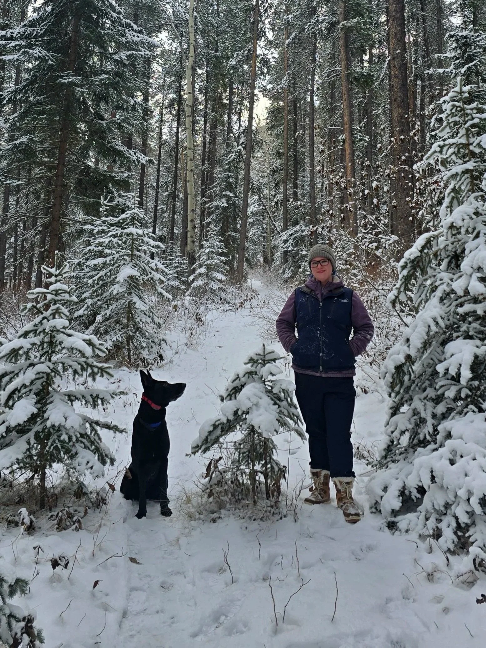 A woman in winter clothing standing on a snowy forest trail next to a black dog, surrounded by snow-covered trees.