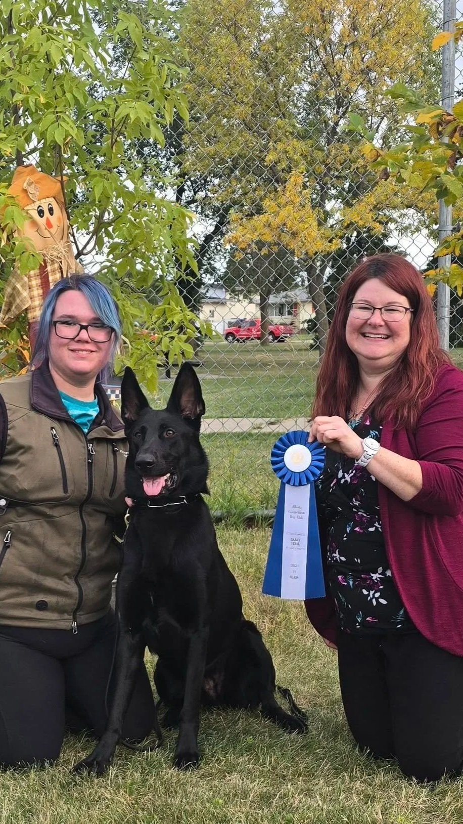 Two women, a dog, and a person in a scarecrow costume outside near a chain-link fence, with trees and parked cars in the background. One woman is holding a blue award ribbon.