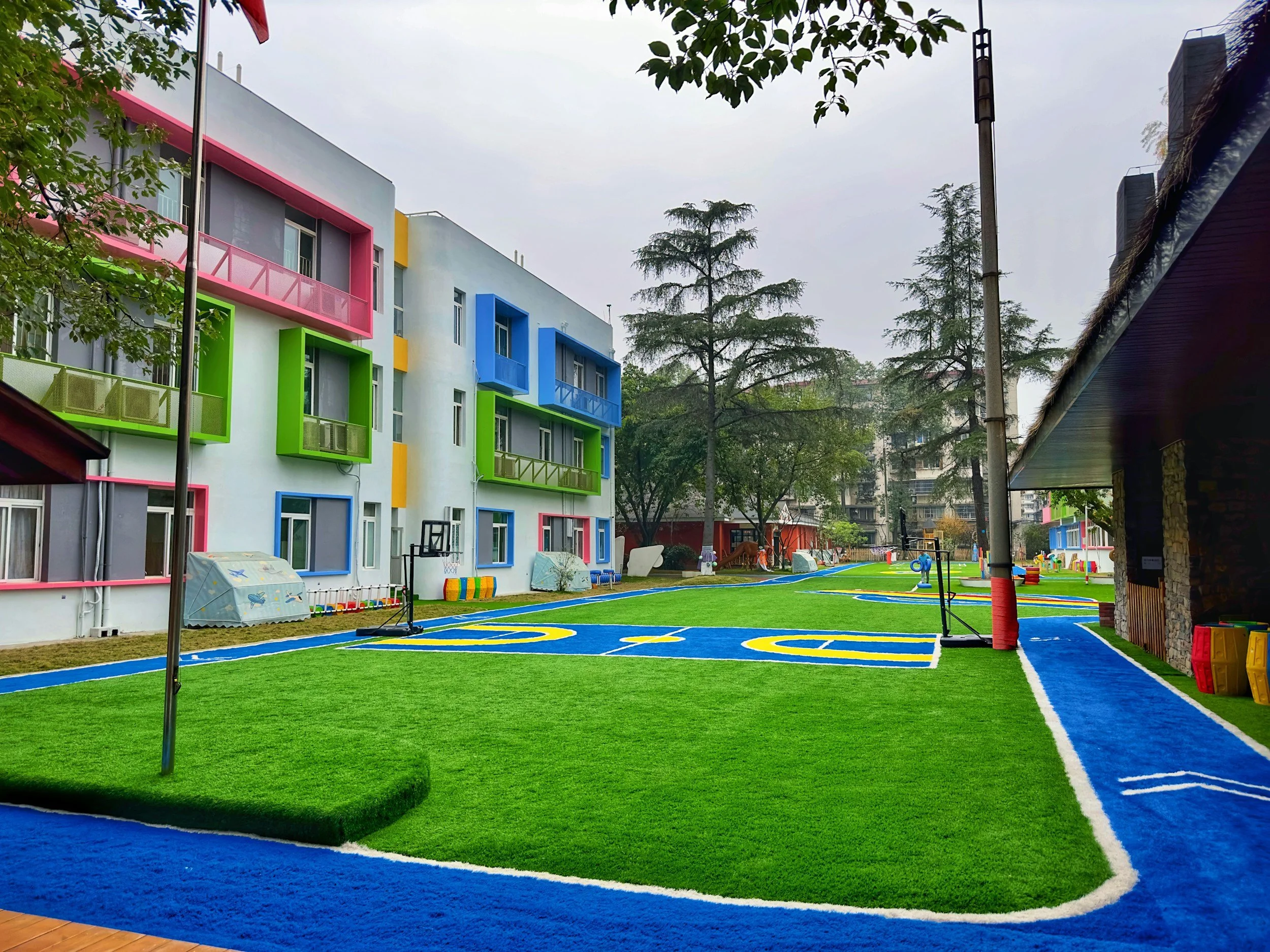 Colorful outdoor playground with synthetic turf, basketball hoops, and multicolored building in the background surrounded by trees.