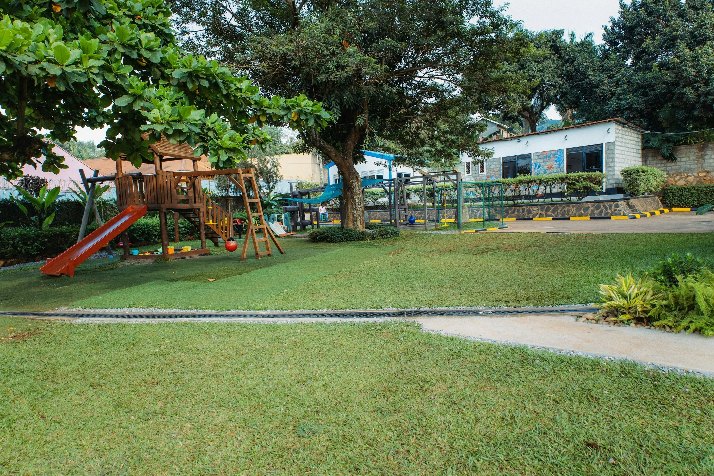 A children's playground with a slide, swings, and a playhouse under a large tree, surrounded by grass, bushes, and trees, with a building in the background.
