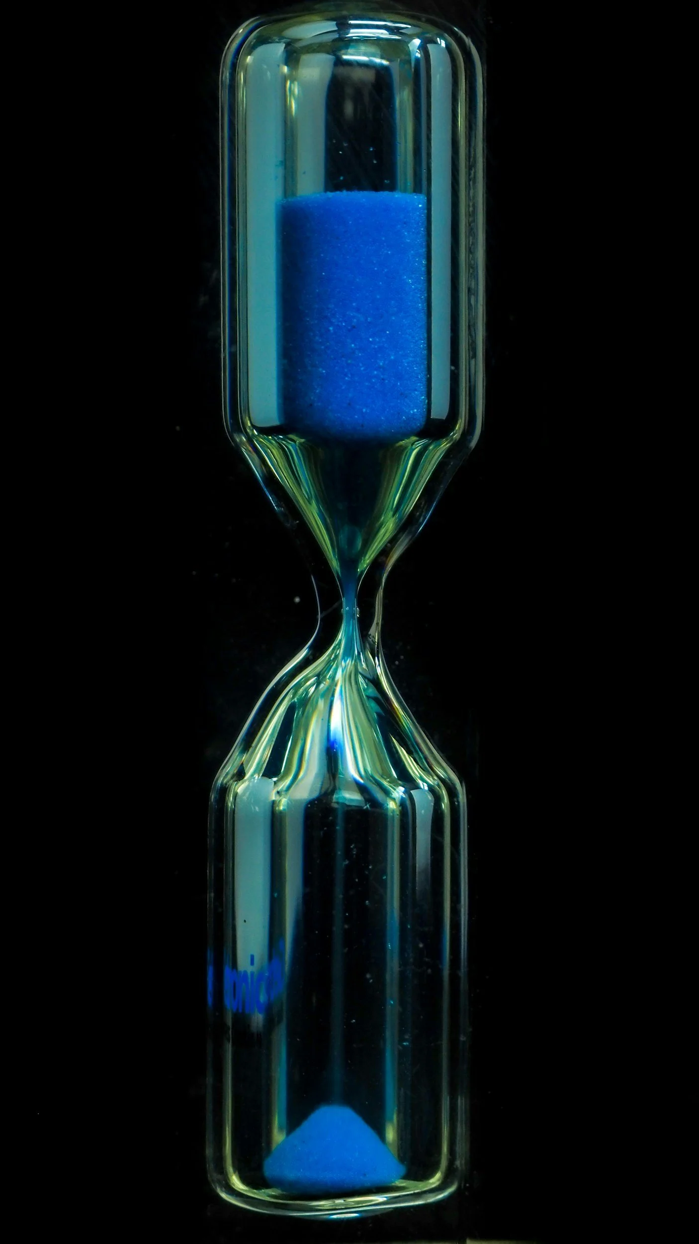 Glass hourglass with blue sand against a black background.