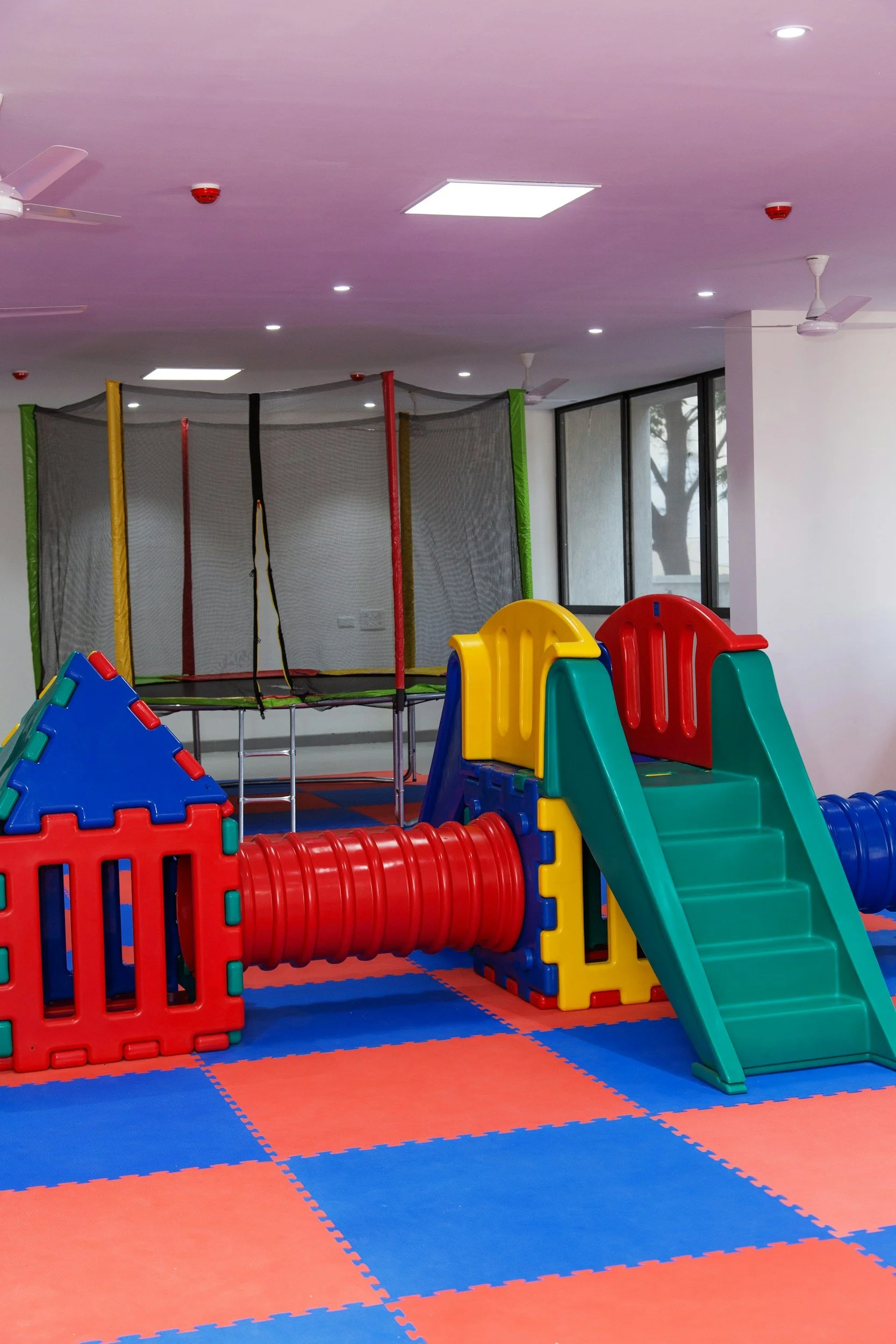 Indoor children's play area with colorful plastic slide, tunnel, and climbing structure, blue and red padded floor, and a trampoline enclosed with safety net.