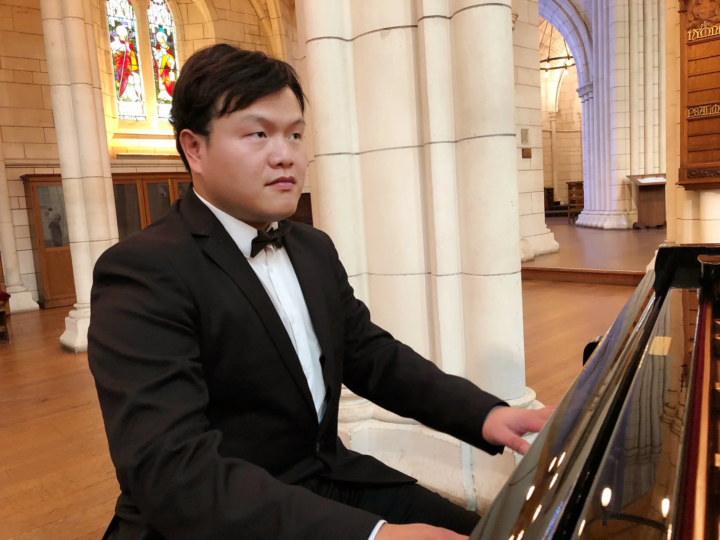 A young man in a black tuxedo playing a grand piano inside a church with stained glass windows.