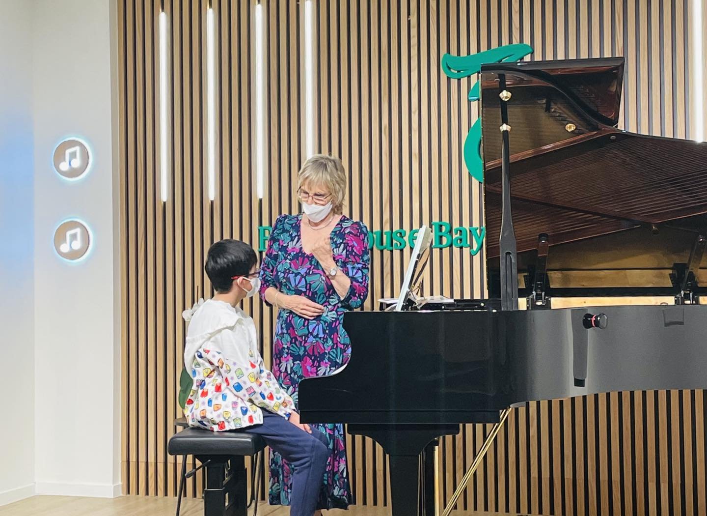 A woman and a young girl, both wearing face masks, are at a black grand piano in a room with a wooden-paneled wall. The woman is standing, wearing a colorful blue and purple dress, and appears to be giving instructions or coaching the girl, who is sitting on a stool, dressed in a white jacket with colorful patterns and dark pants.
