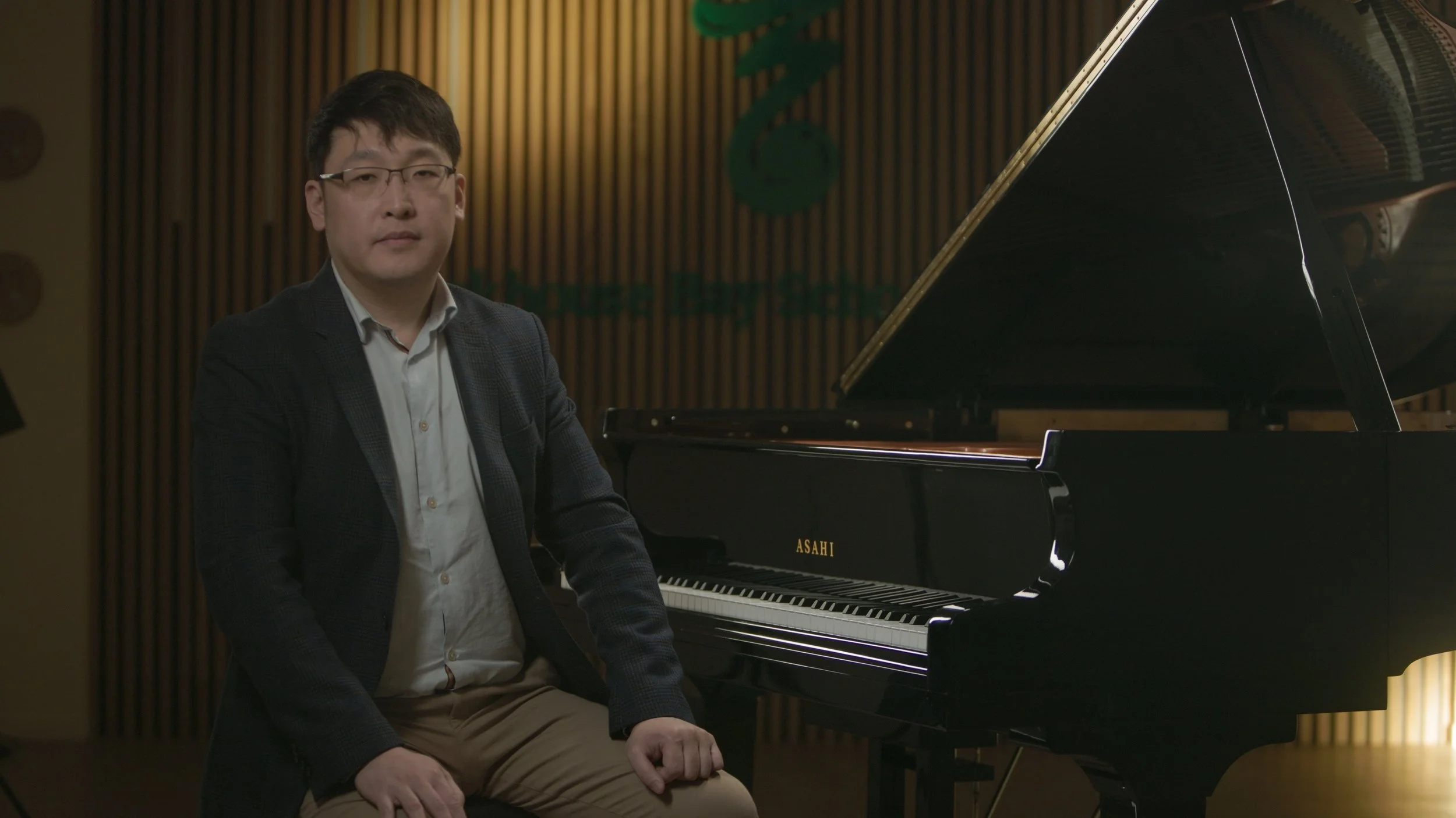 A man wearing glasses, a light shirt, and a dark blazer sitting beside a black grand piano in a music studio with wooden wall panels and a green logo.