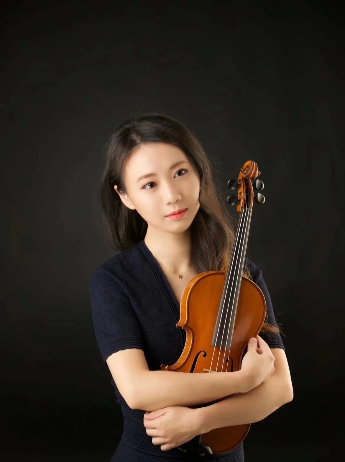 Young woman with long dark hair holding a violin against a black background.