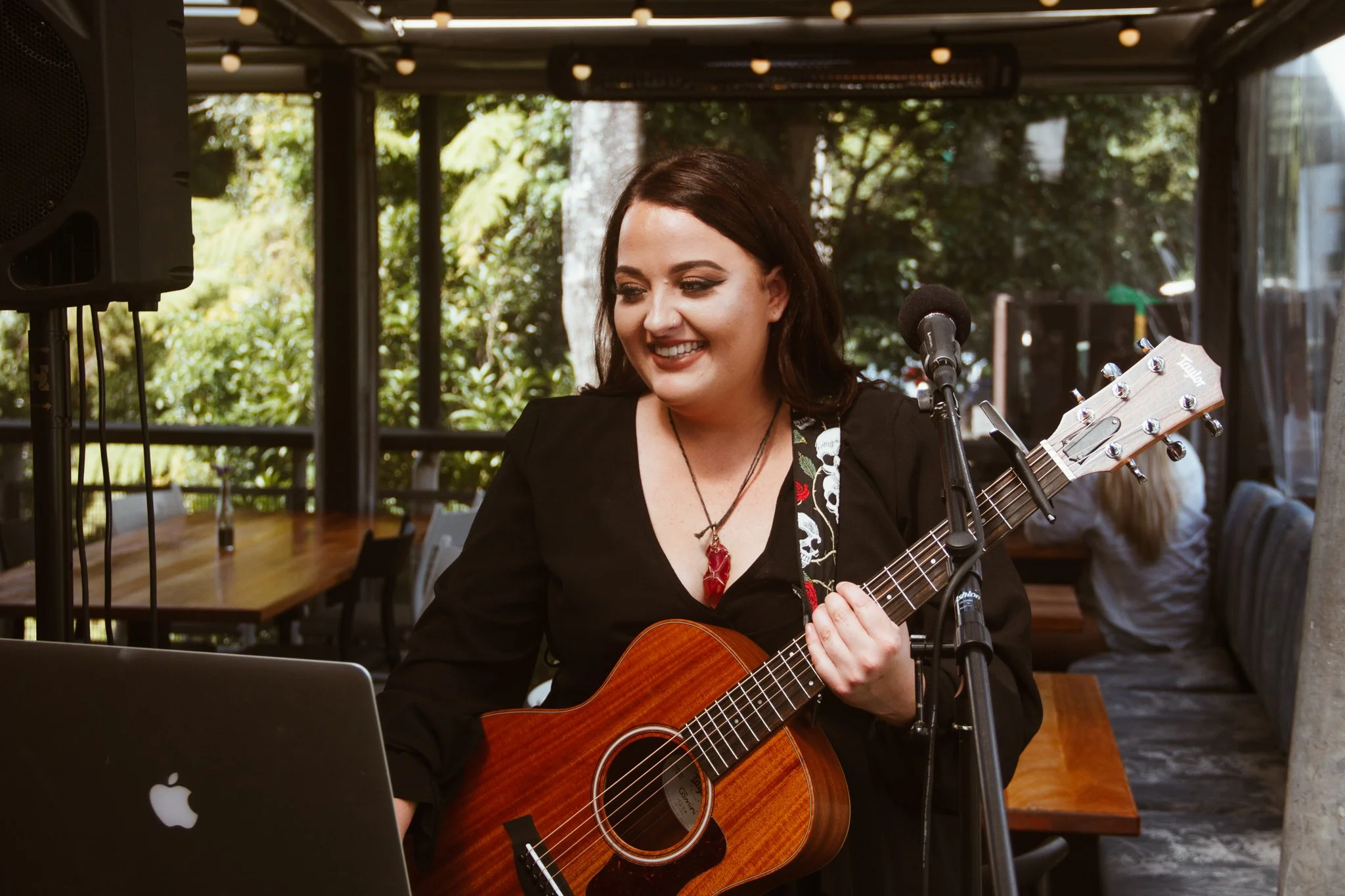 A woman with dark brown hair smiling while playing an acoustic guitar and using a laptop at an outdoor venue with trees in the background.