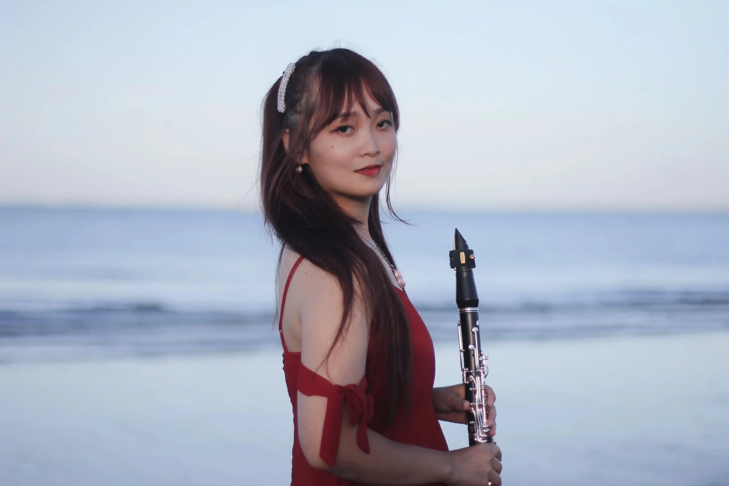 Woman with long dark hair in a red dress holding a clarinet on a beach with calm water and a light sky.