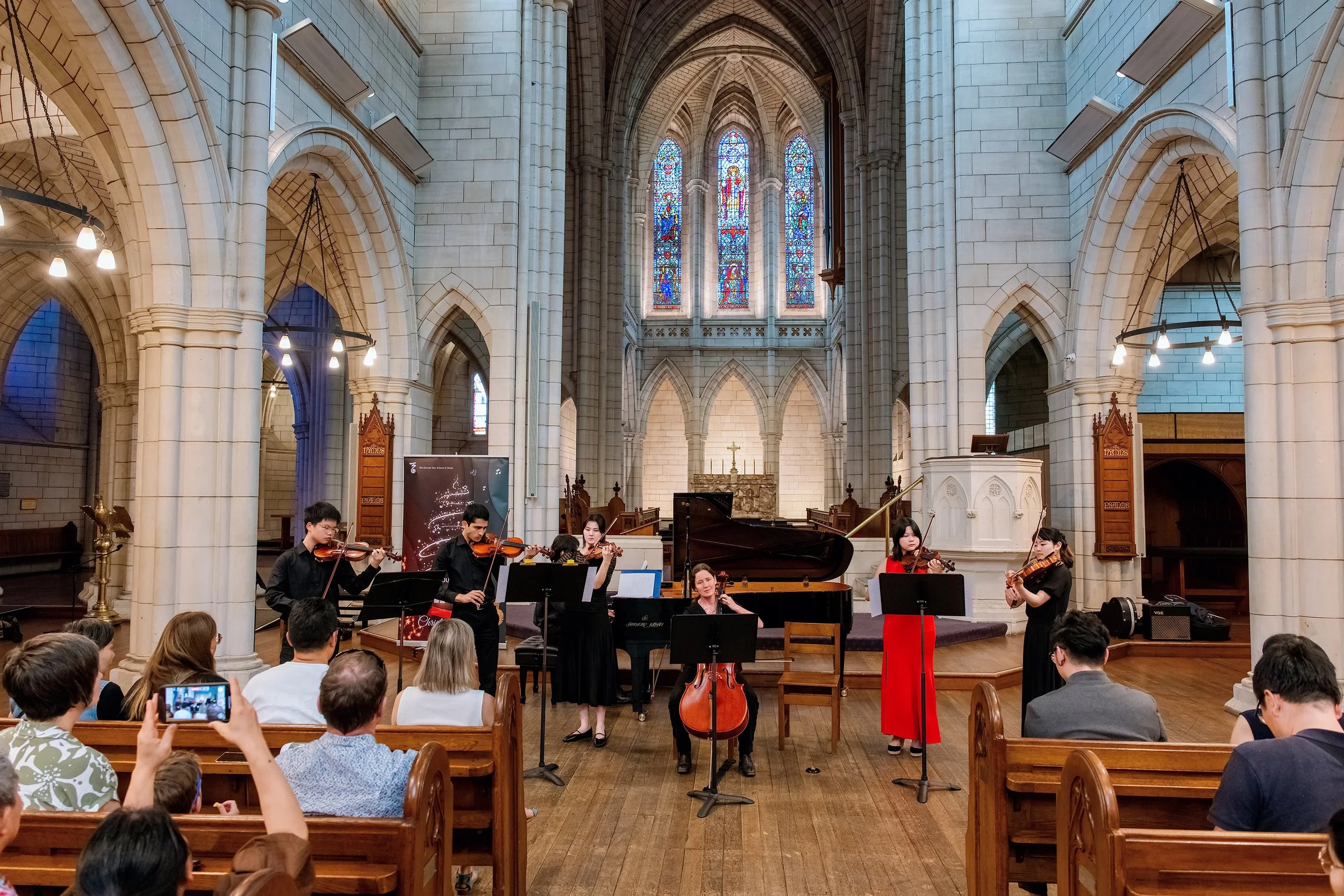 A small orchestra performing inside a grand Gothic-style church with stained glass windows, and an audience seated and watching.