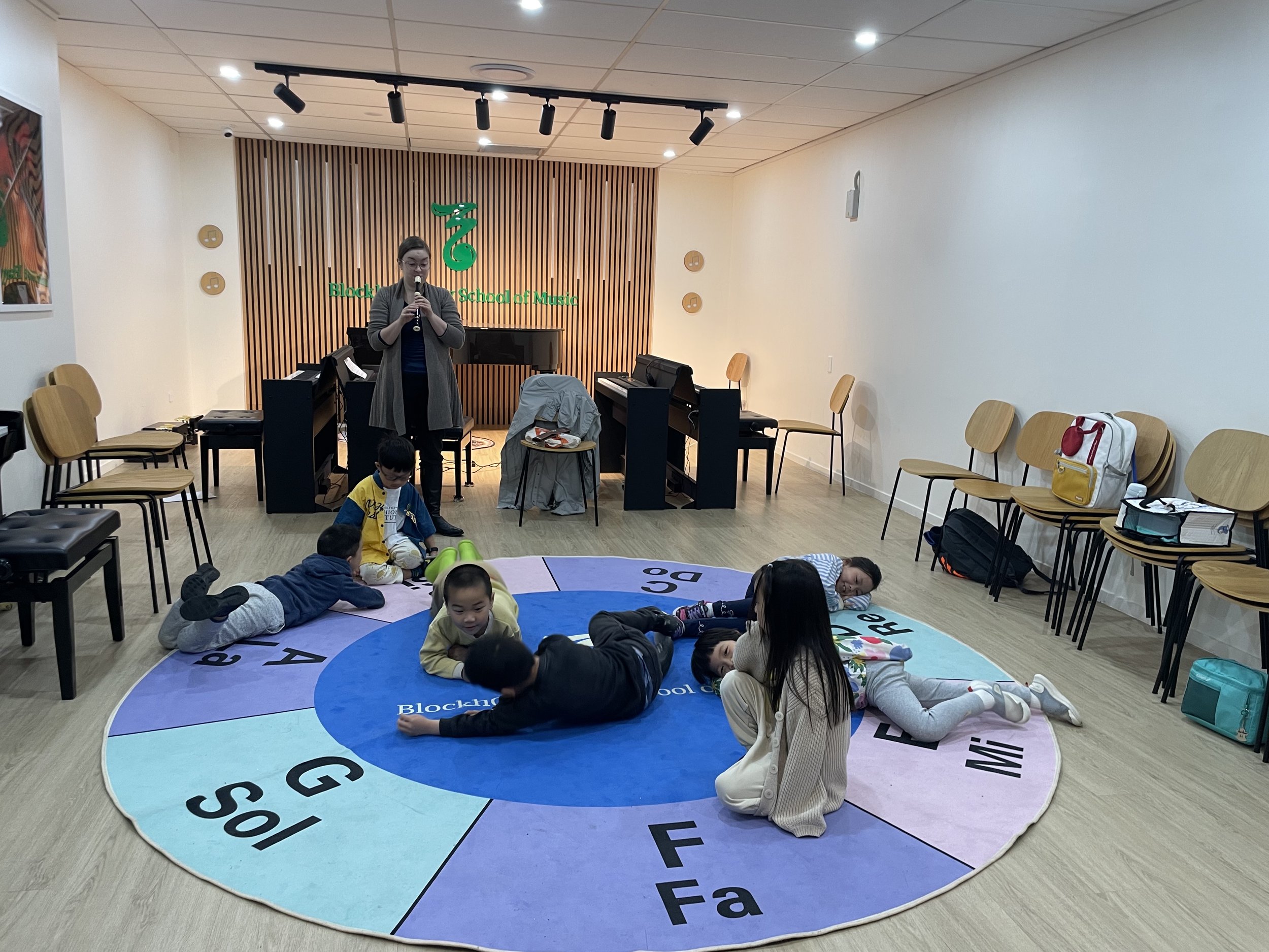 Children playing on a colorful circular rug in a music classroom, with a woman singing into a microphone at the front. Chairs are lined up along the walls, and musical instruments and equipment are visible.
