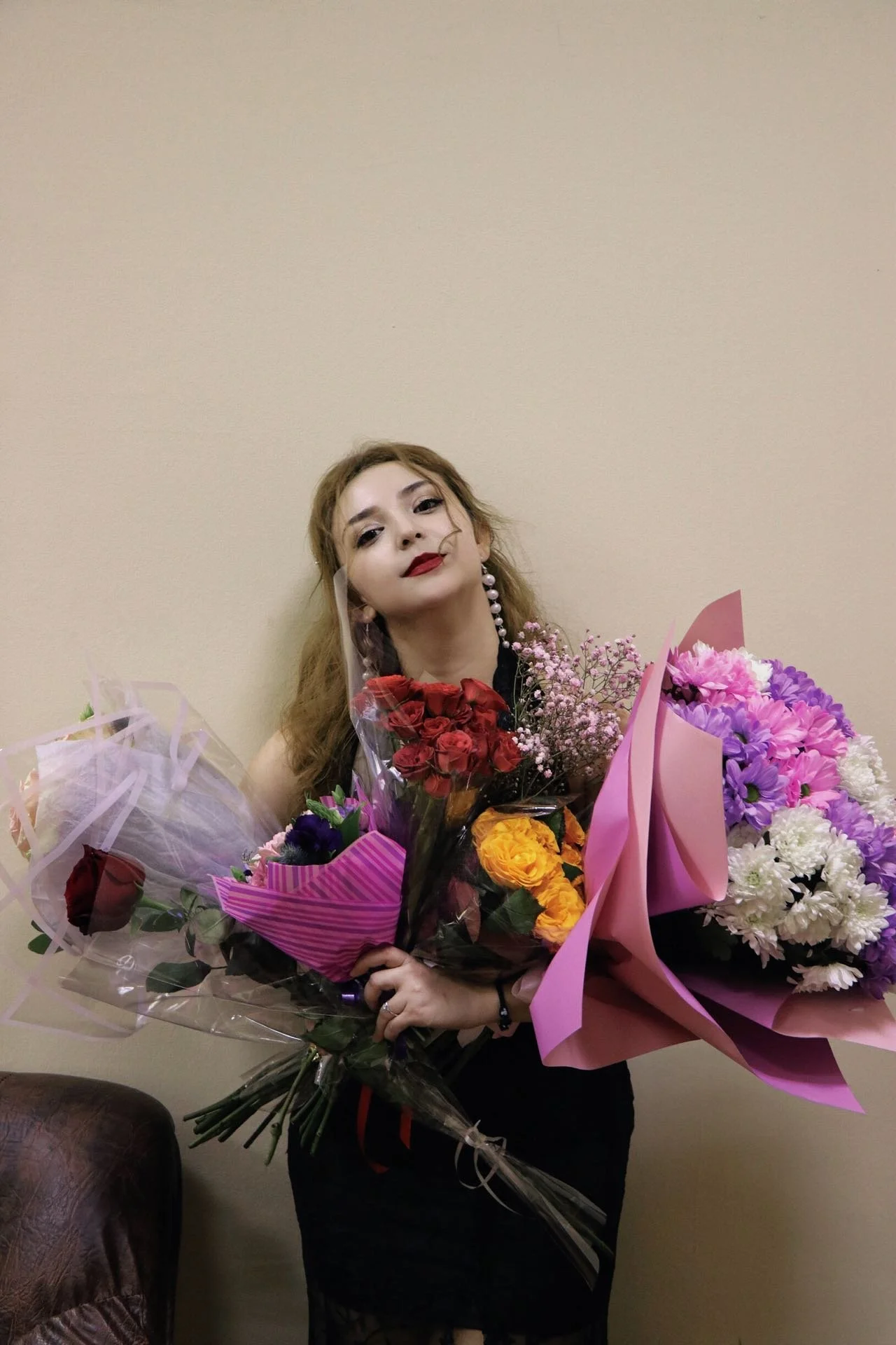 A woman with wavy hair, wearing makeup with red lipstick, holding multiple bouquets of colorful flowers, standing against a plain beige wall.