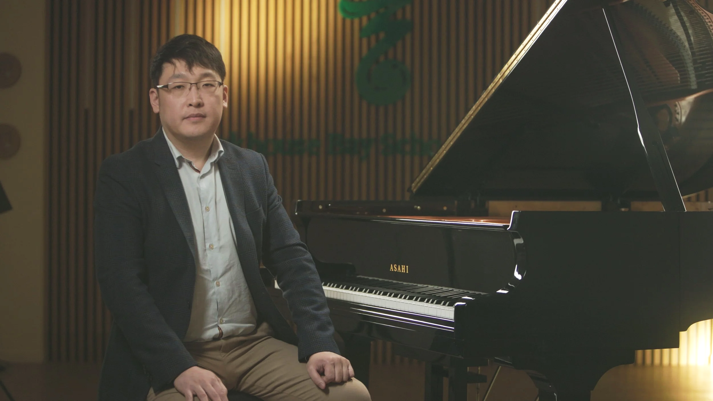 Man sitting next to a black grand piano in a room with wooden wall panels.