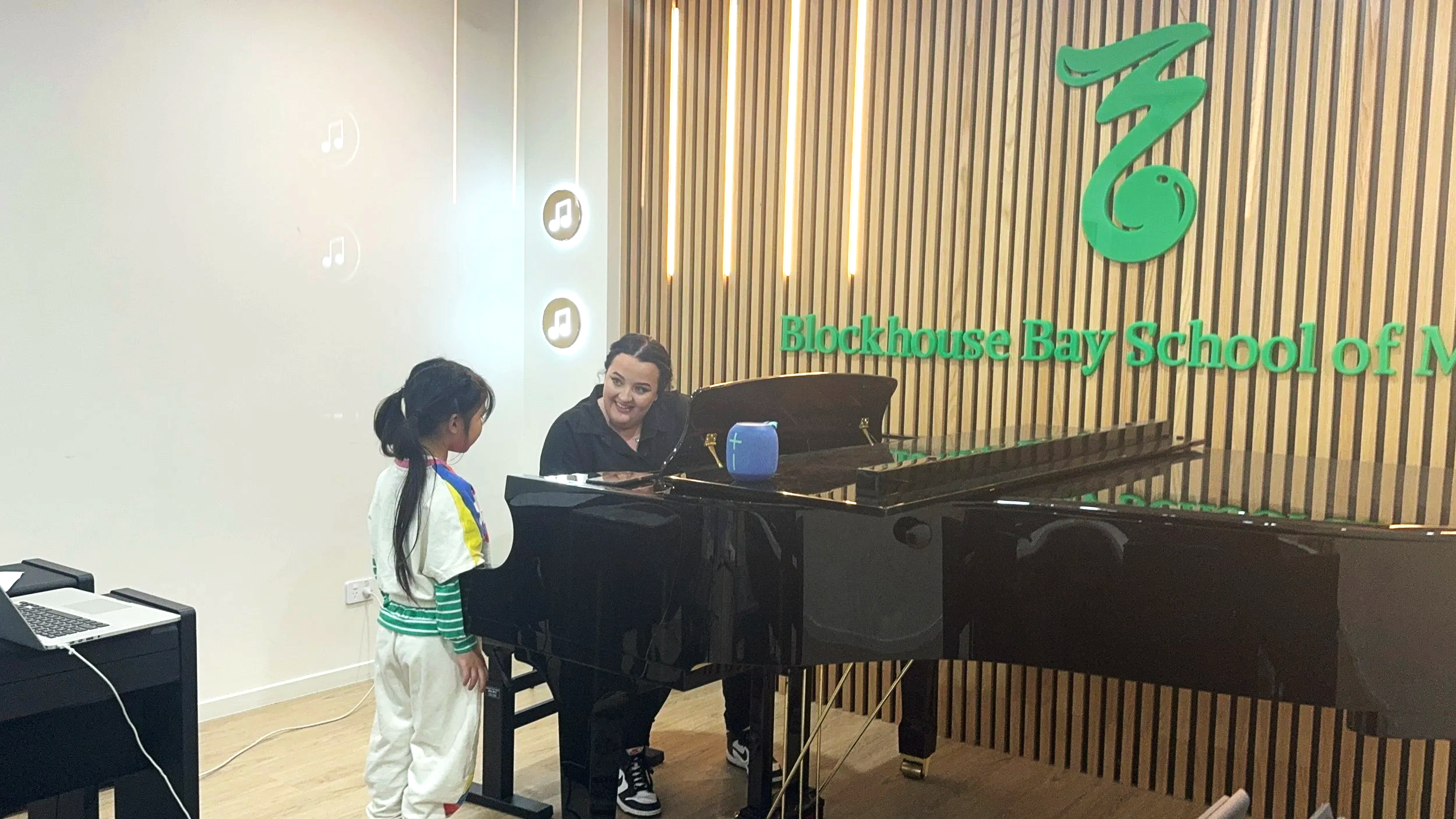 A woman playing a black grand piano and a young girl in a colorful outfit standing next to her in a music classroom at Blockhouse Bay School of Music.