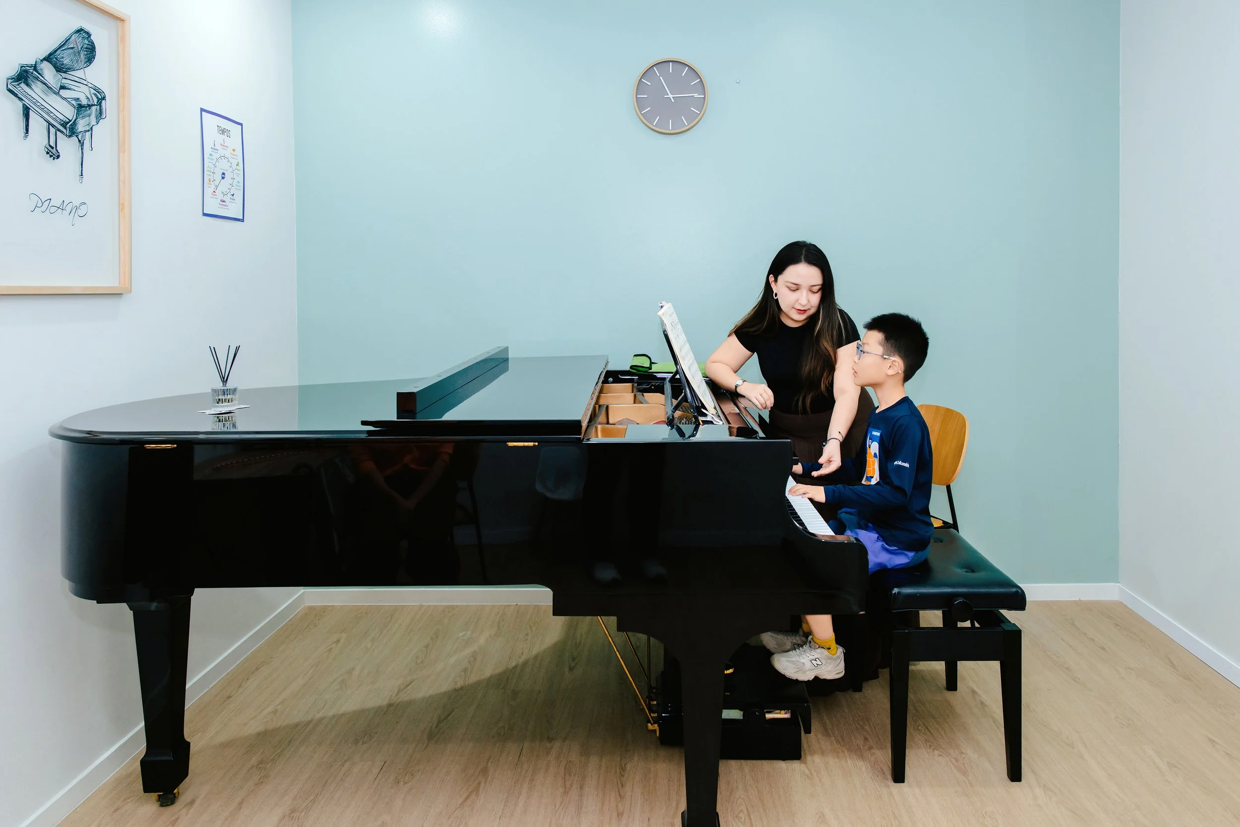 A music teacher instructing a young boy at a grand piano in a classroom with light blue walls and a wall clock.