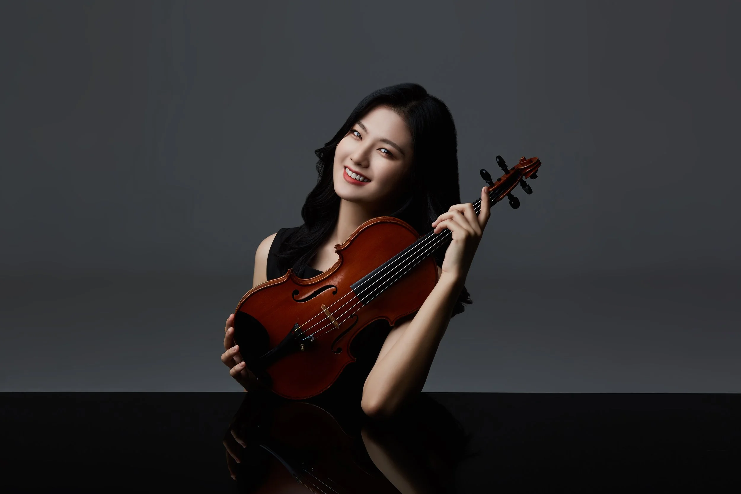 A smiling woman with dark hair holding a violin in front of a dark background.