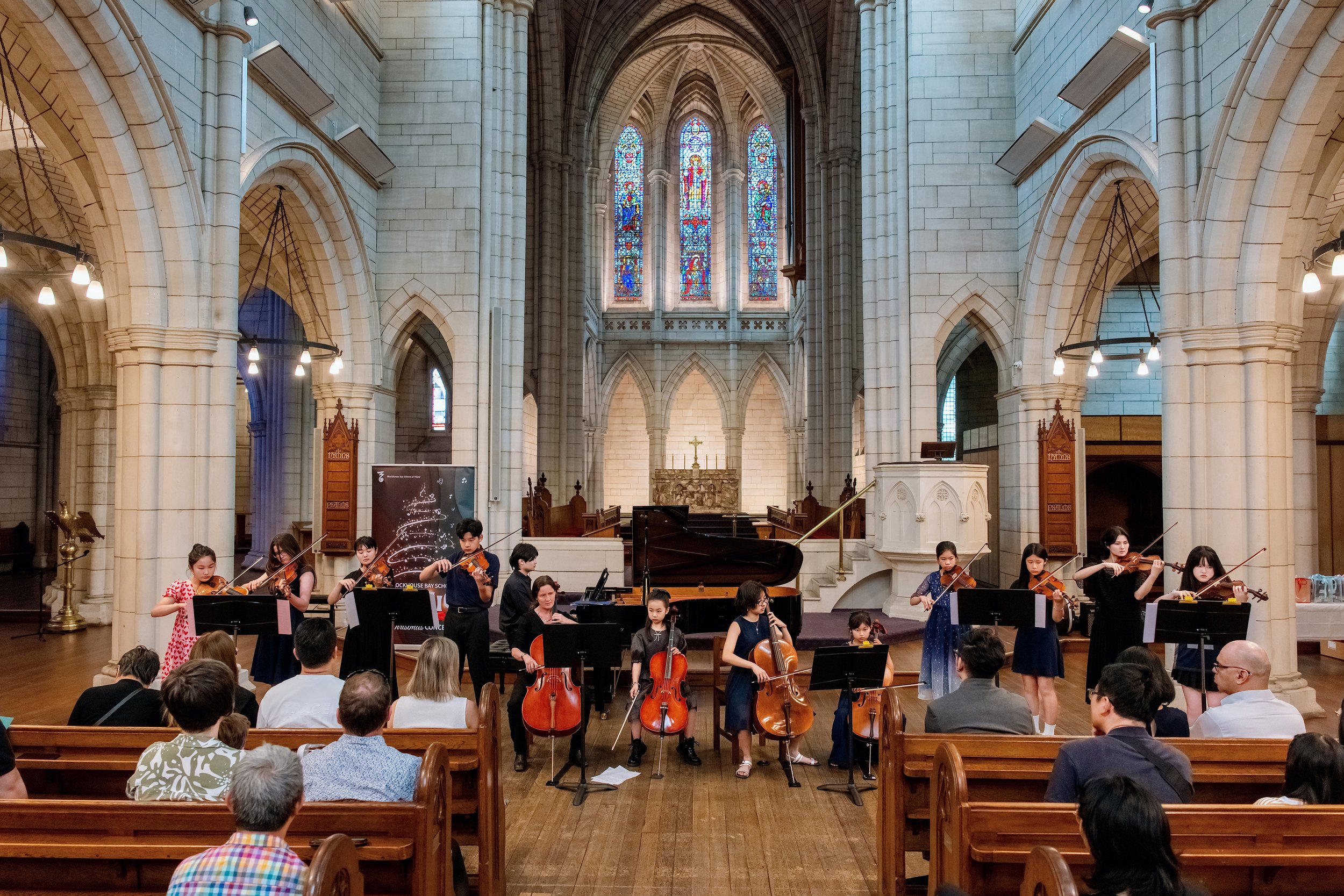 Children playing violins, cellos, and piano during a concert inside a large, ornate church with stained glass windows.
