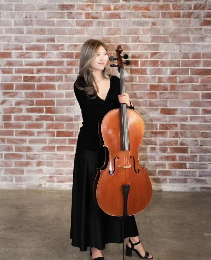 Woman in black dress holding a cello in front of a brick wall.