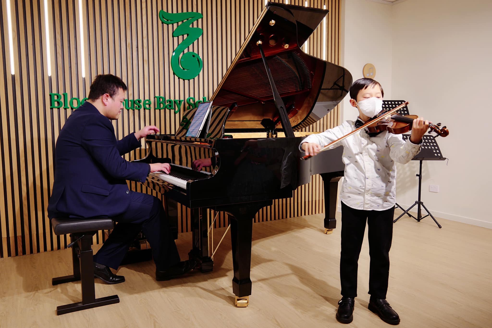 A piano and violin performers in a performance hall. The pianist is wearing a dark suit, and the violinist is a child wearing a white shirt and a face mask. The background has a wooden panel wall with green signage that reads 'Bloom Use Bay School'.