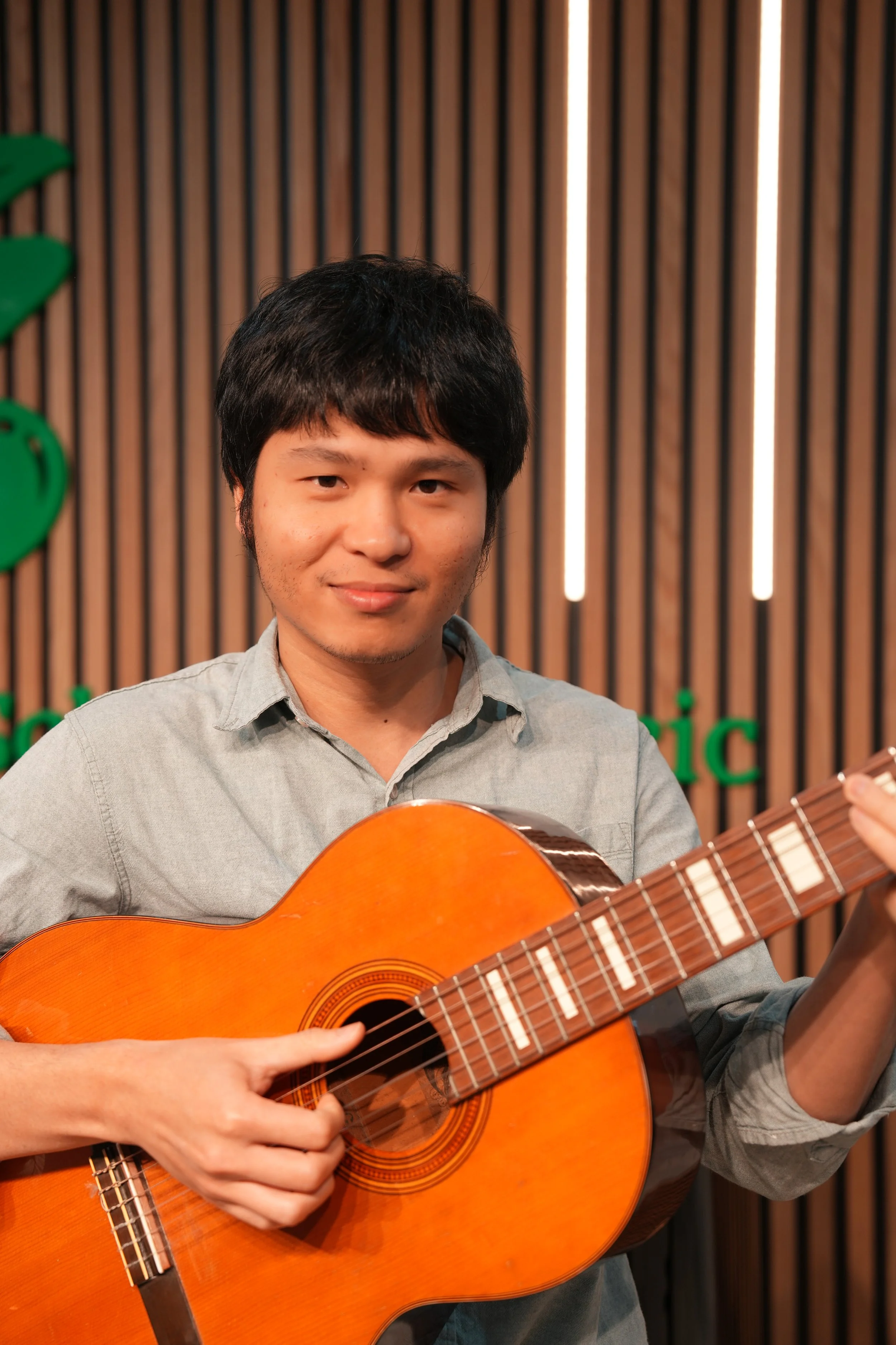A young man playing an orange acoustic guitar indoors with a green and brown wooden wall behind him.