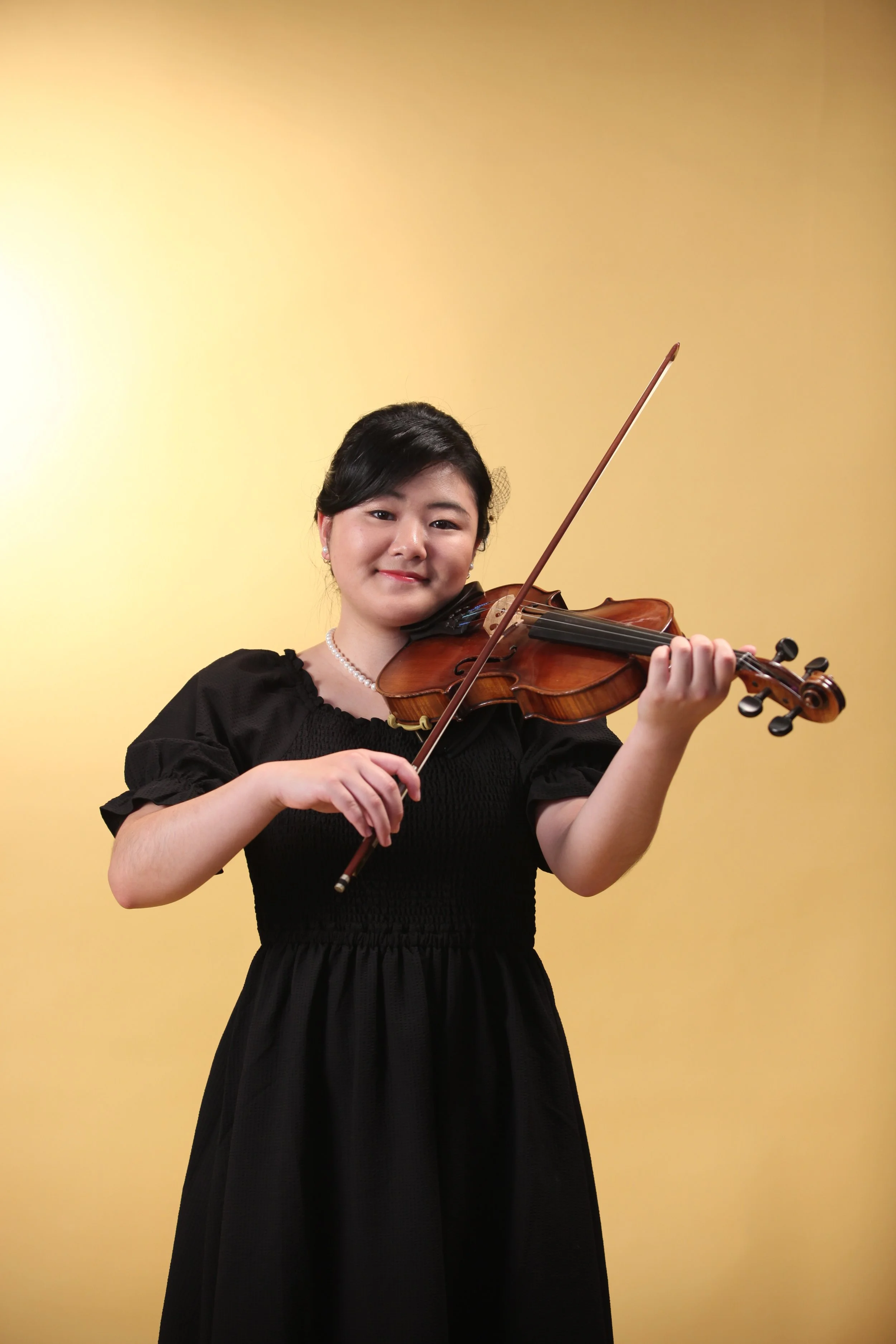 Young woman with black hair, wearing a black dress, playing a violin against a yellow background.