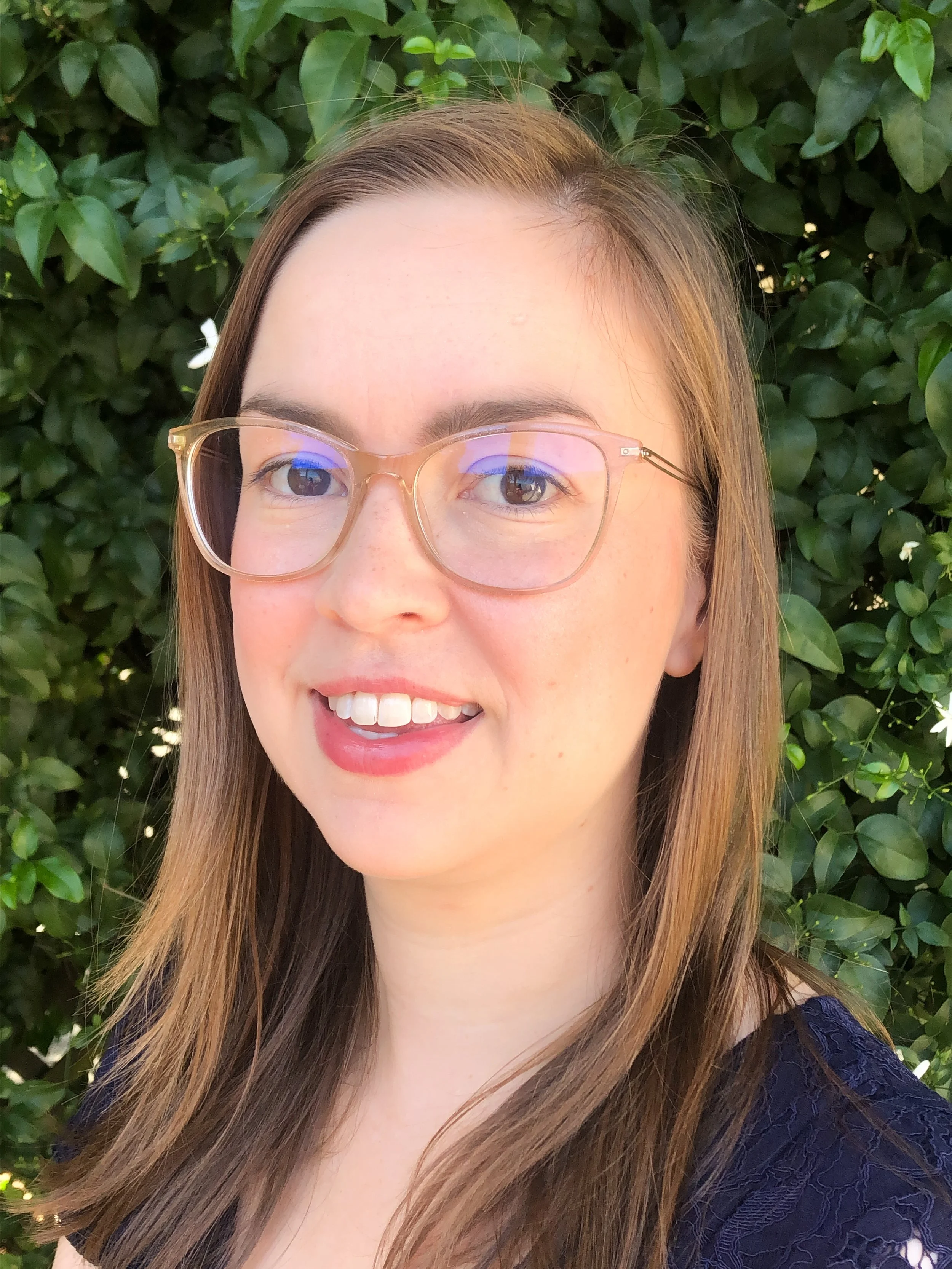 A young woman with long light brown hair, wearing glasses with a light gold frame, smiling, standing outdoors in front of a green leafy background.