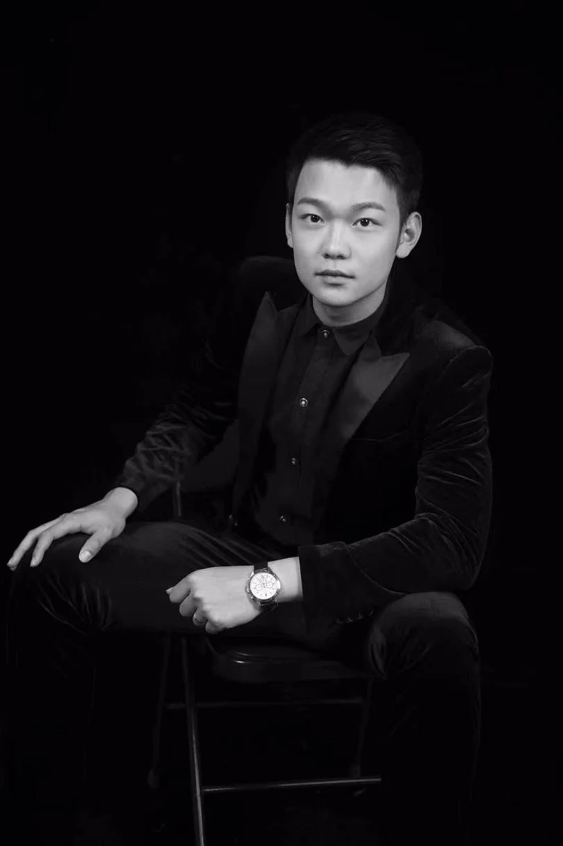 A young man dressed in a black suit and shirt, seated on a chair against a dark background, looking at the camera with a neutral expression, wearing a watch on his left wrist.