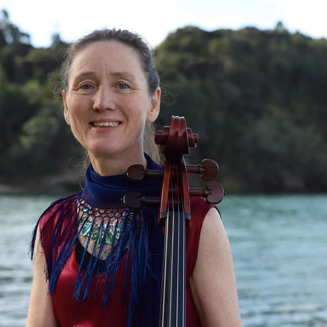 A woman with light skin and blue eyes smiling, holding a cello outdoors with water and trees in the background.